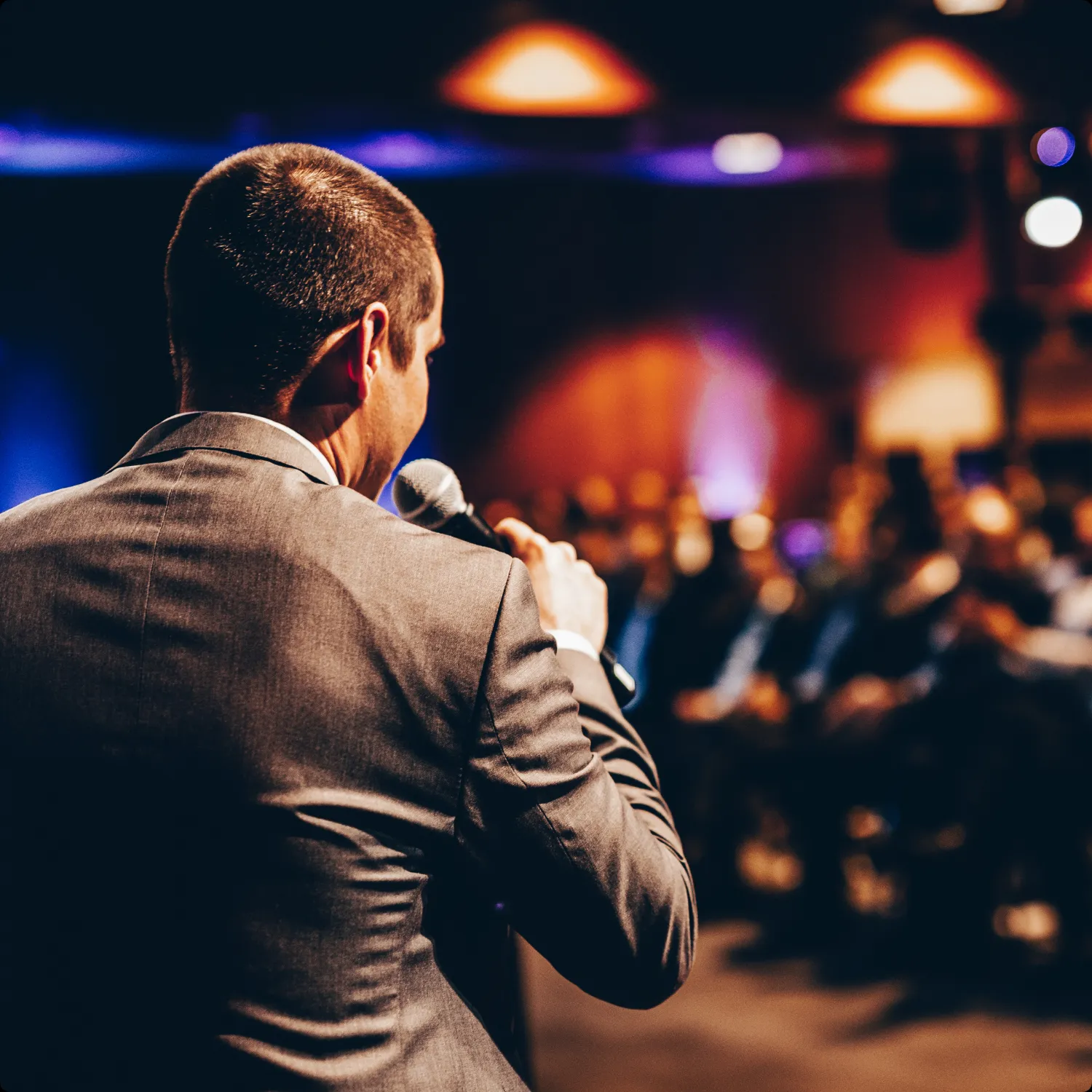 Man in suit holding microphone and speaking to an audience in a dimly lit event space.