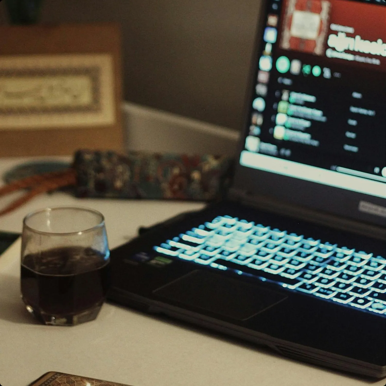 Glass of dark beverage next to a laptop with illuminated keyboard on a desk.