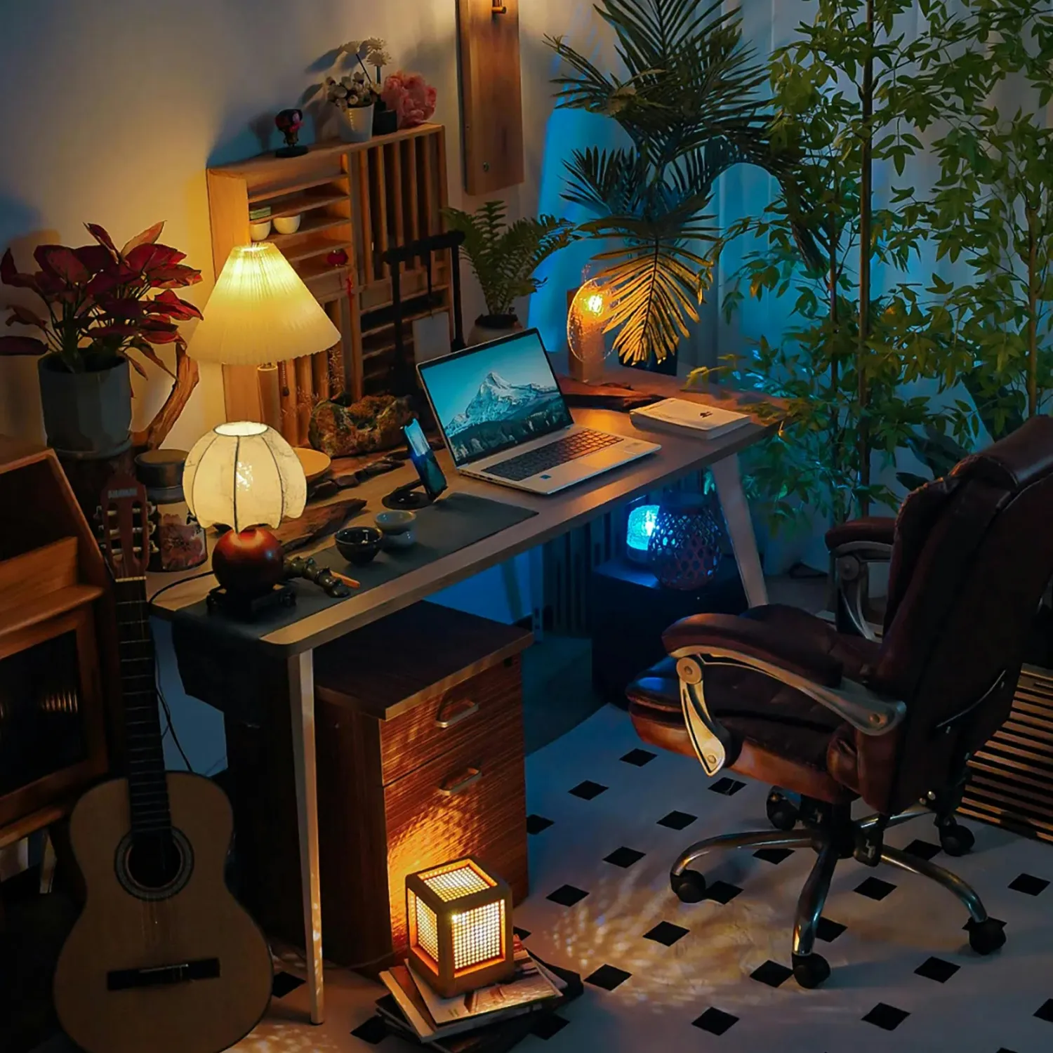 Cozy home office setup with a desk holding a laptop, smartphone, two lit lamps, various plants, and a brown swivel chair on a black-and-white tiled floor.