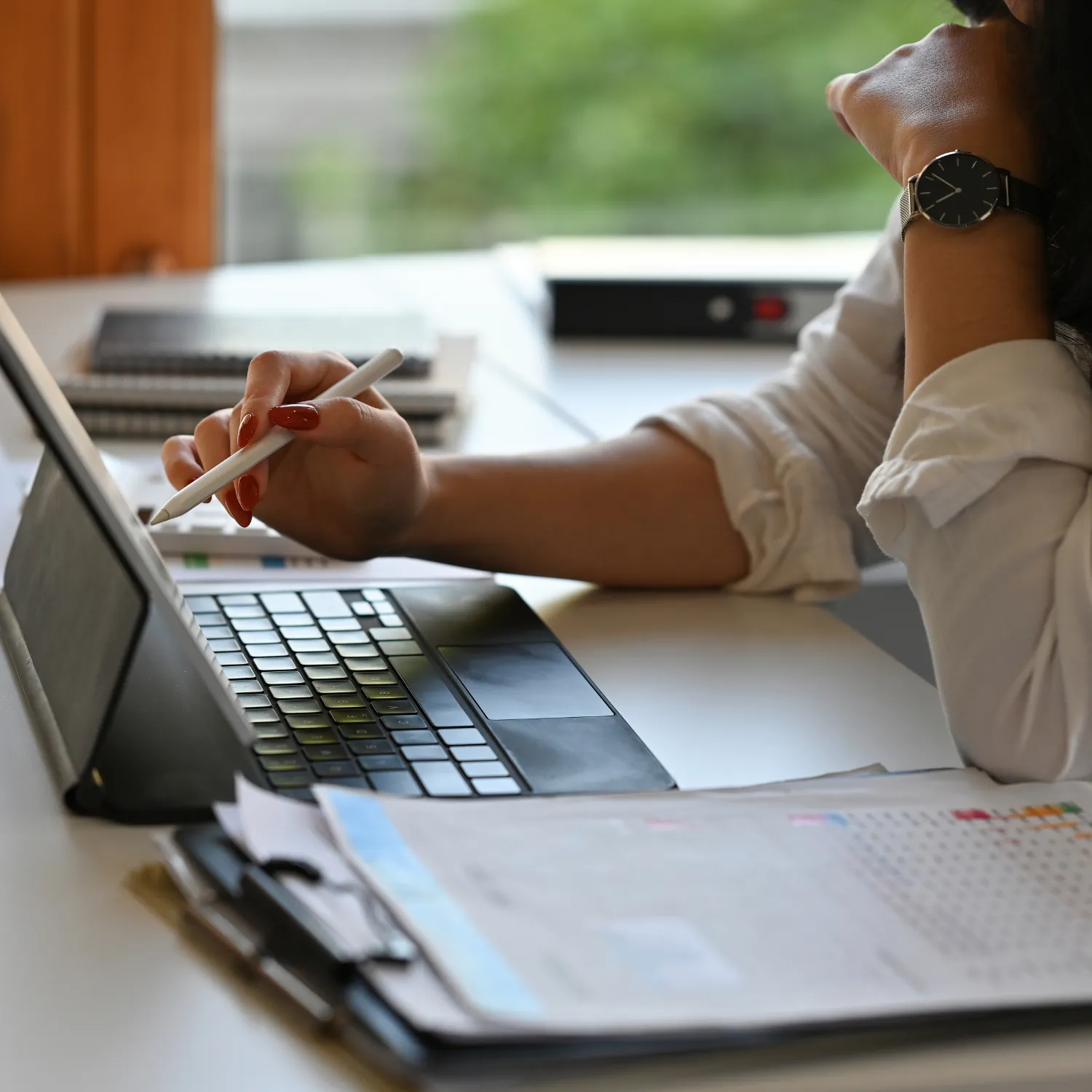 Person using a stylus pen on a tablet with a keyboard, next to documents and notebooks on a desk.