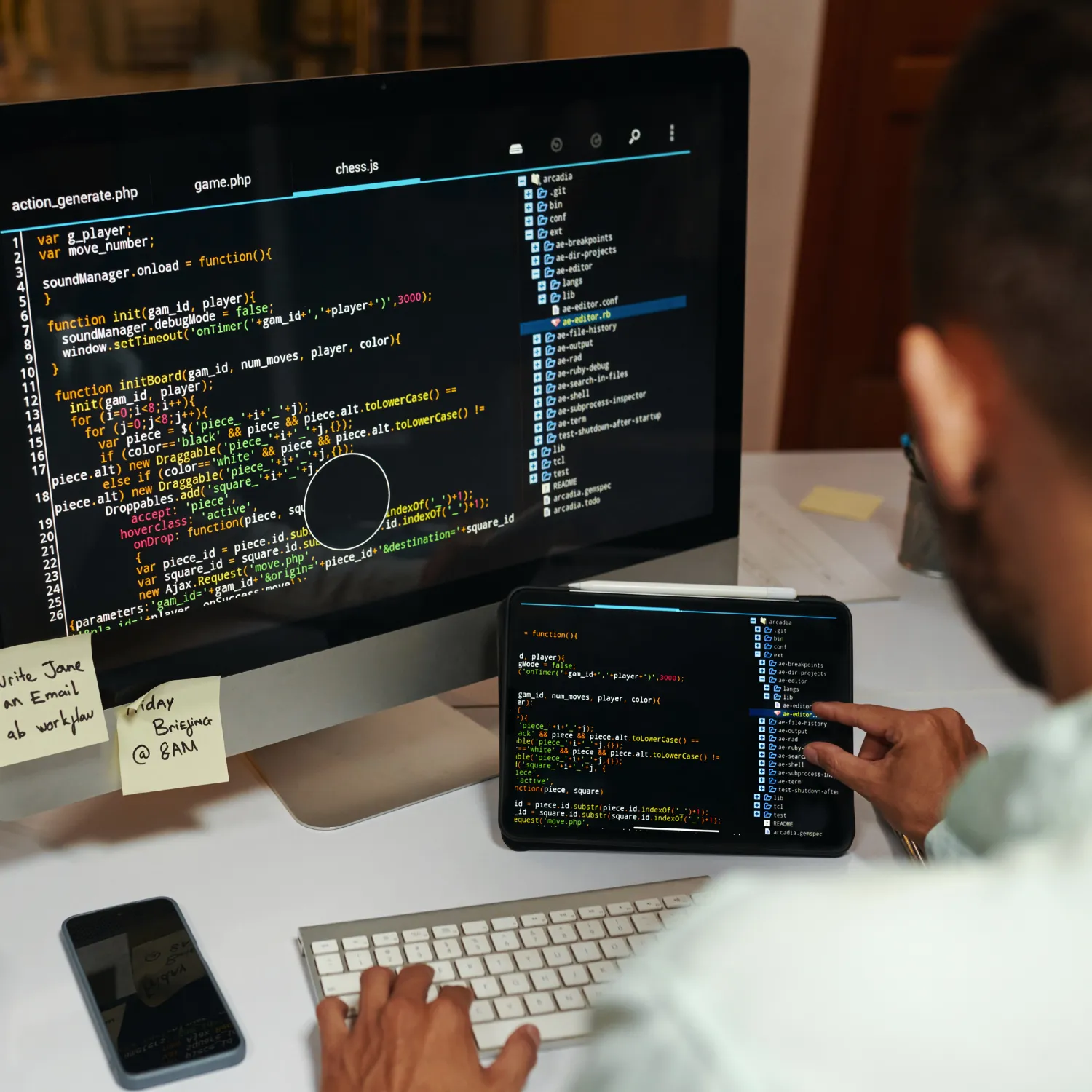 Person working on JavaScript code displayed on both a desktop monitor and a tablet at a white desk with a keyboard and smartphone.