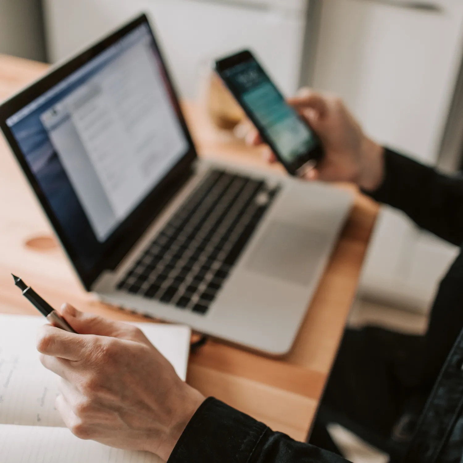 Person holding a fountain pen and writing in a notebook next to a laptop and smartphone on a wooden desk.