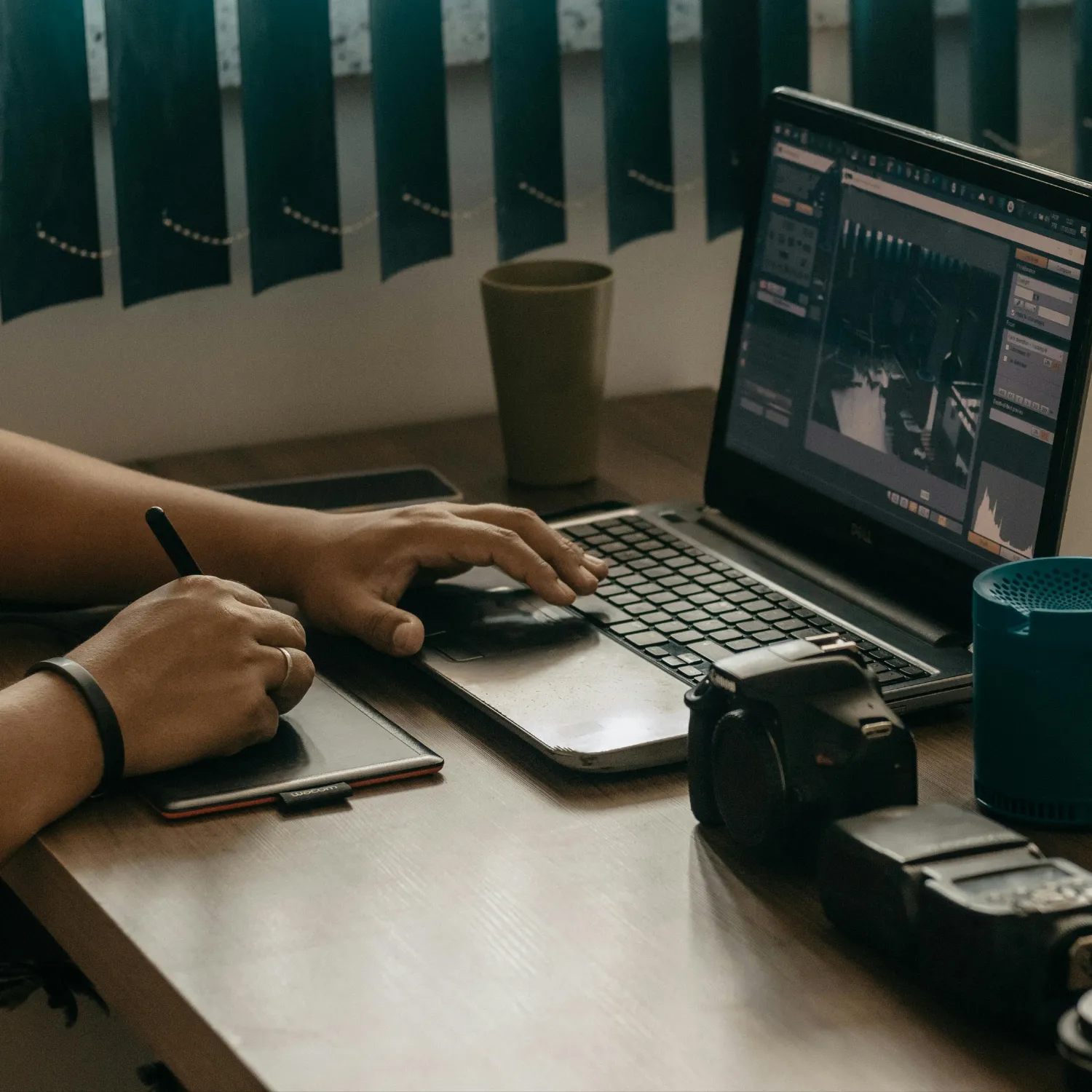 Person using a Wacom tablet with a stylus and editing photos on a laptop screen, with a camera and cup nearby on the desk.