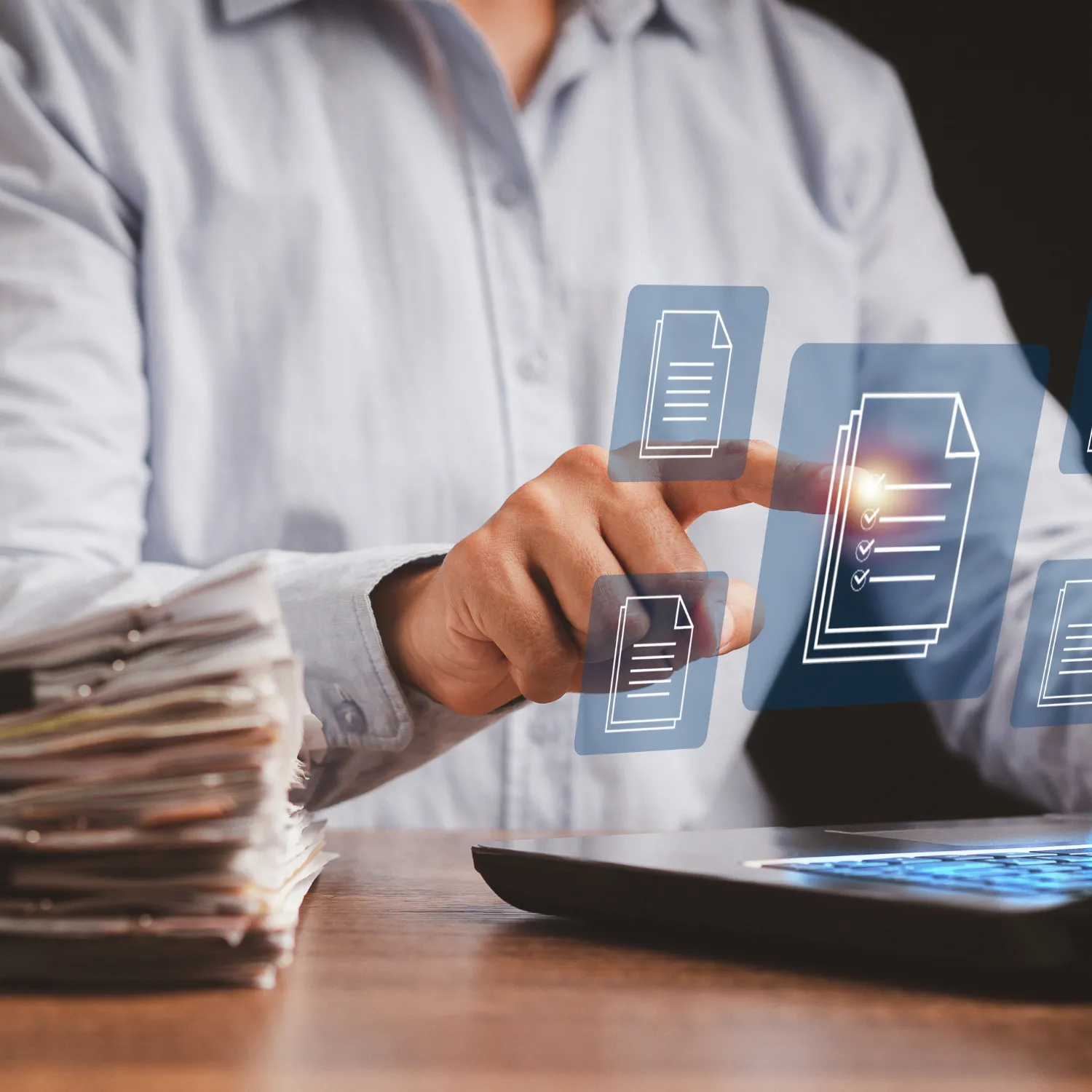 Person interacting with digital document icons above a laptop keyboard, with a stack of paper documents beside them.