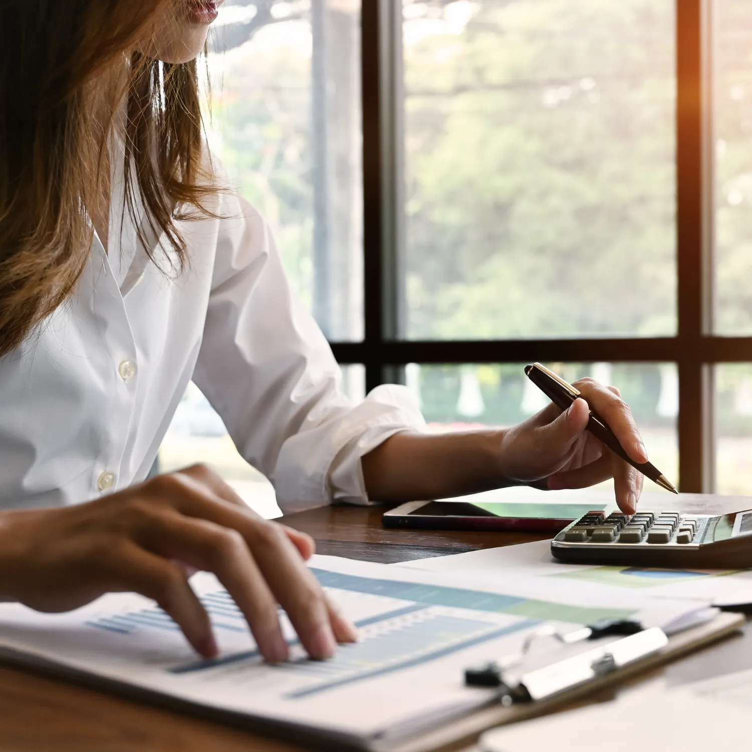 Person in white shirt using a calculator and reviewing financial documents on a clipboard by a window.