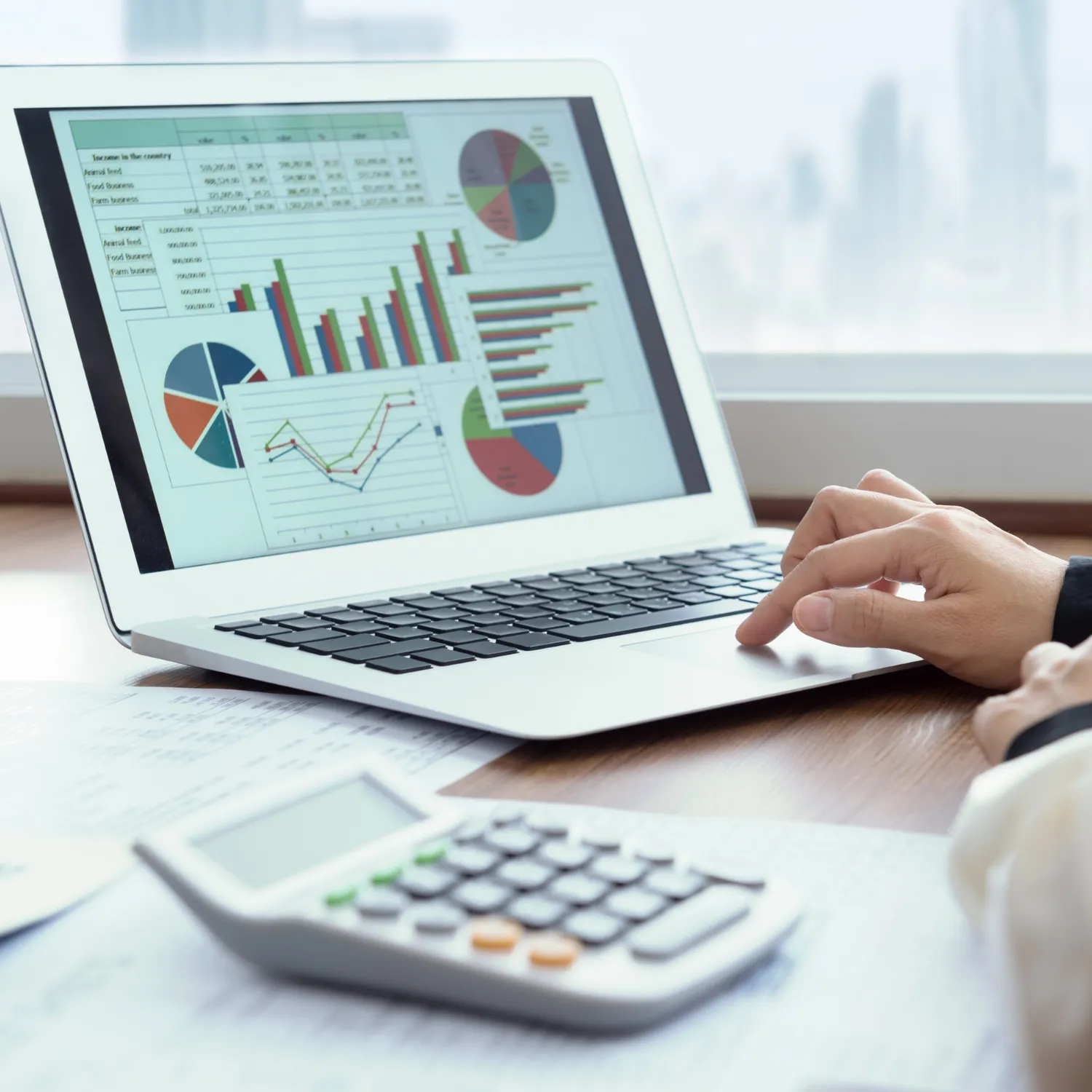Person using a laptop displaying various colorful financial charts and graphs with a calculator in the foreground on a wooden desk.