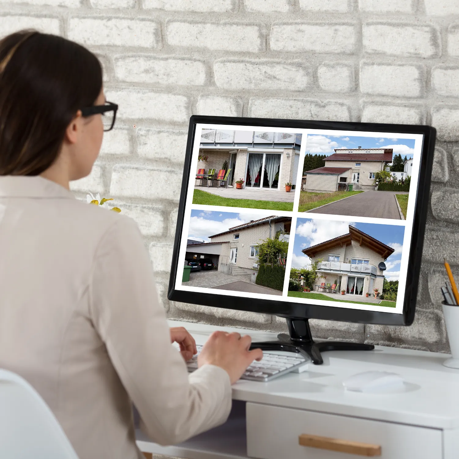 Woman with glasses sitting at a desk looking at a computer screen displaying four outdoor photos of a beige brick house.