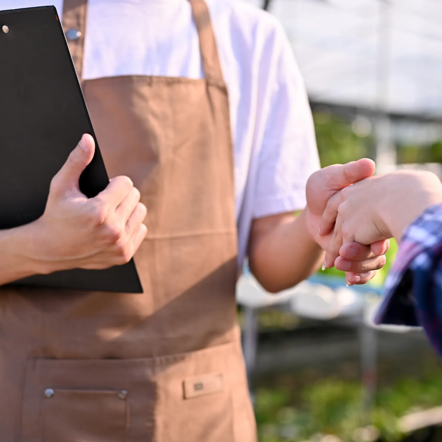 Person in brown apron holding a clipboard shaking hands with another person outdoors.
