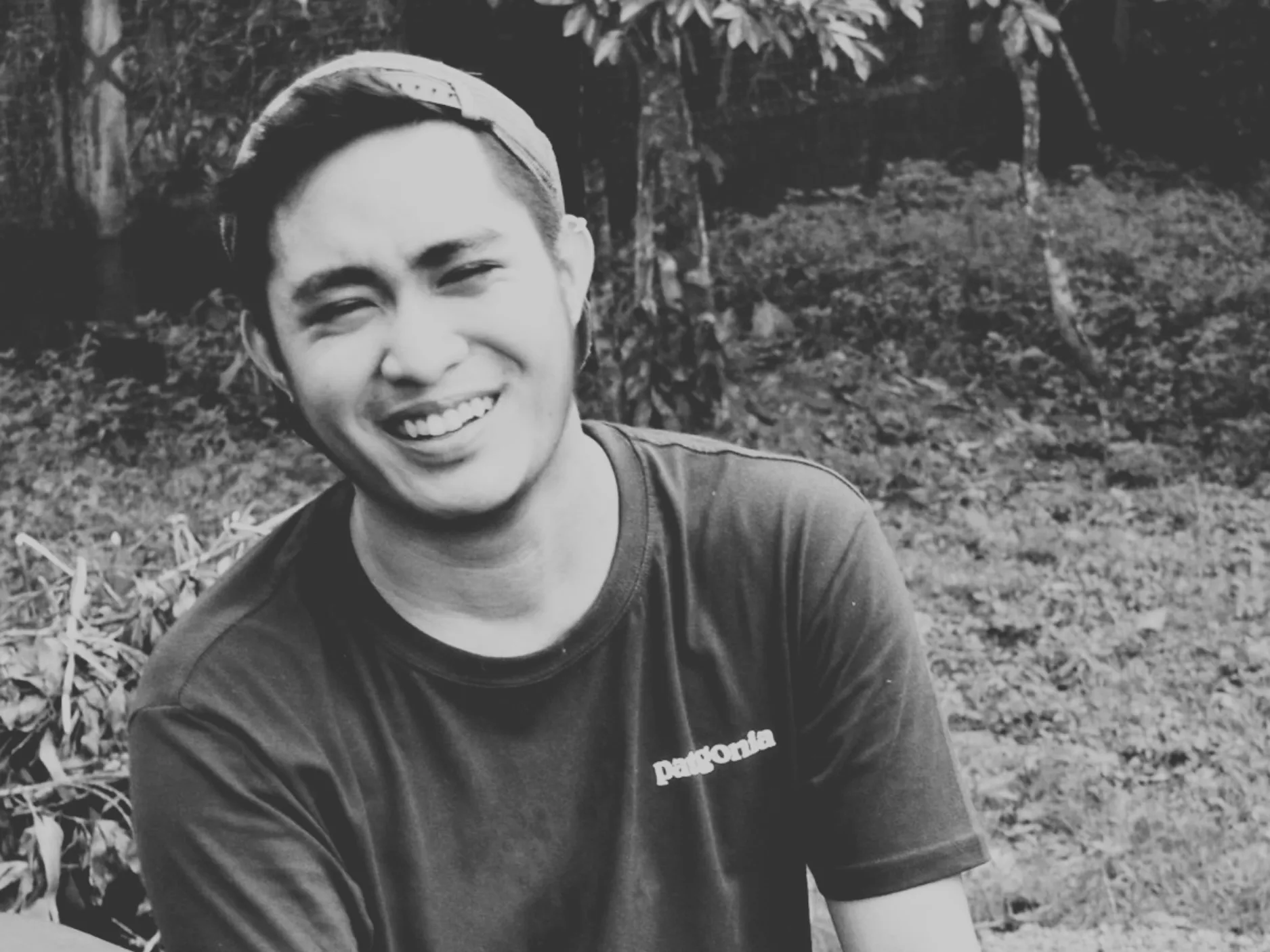 Young man wearing a cap backwards, smiling outdoors with trees and grass in the background.