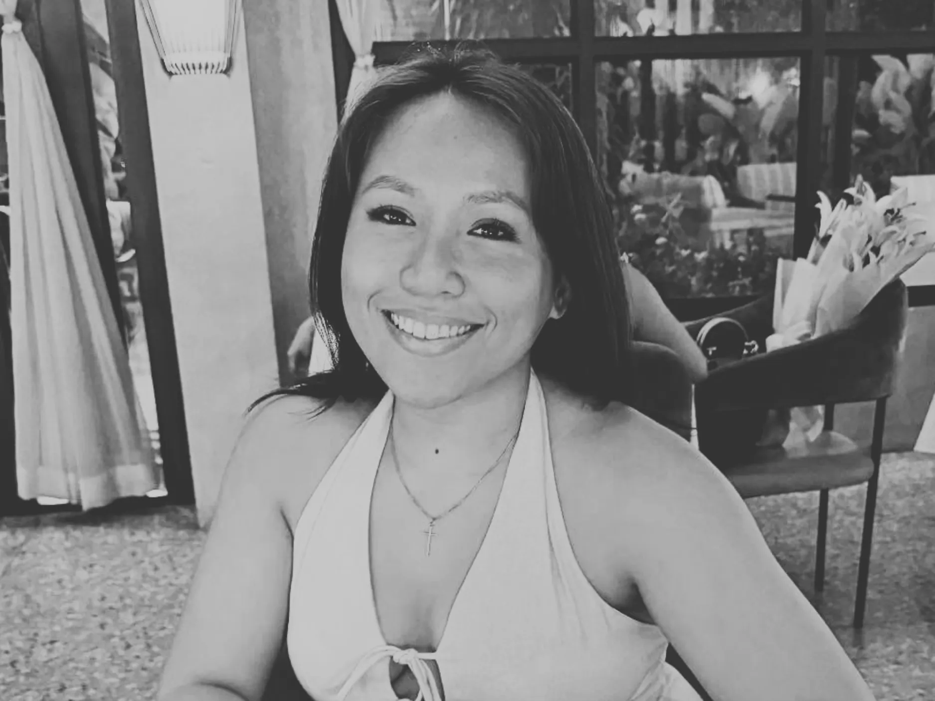 Smiling woman with long hair wearing a halter top and a cross necklace, sitting indoors near a window.