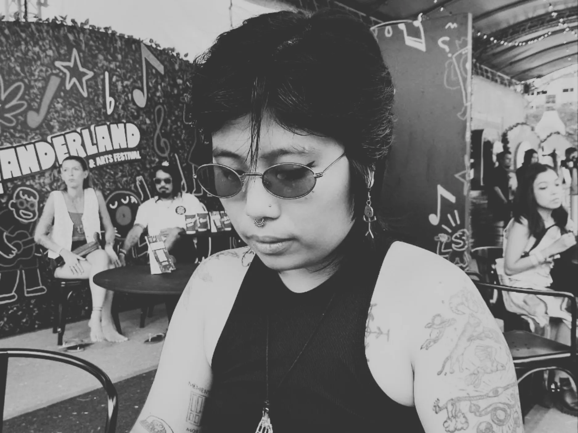 Woman with tattoos, nose ring, and sunglasses looking down at an outdoor arts festival seating area.