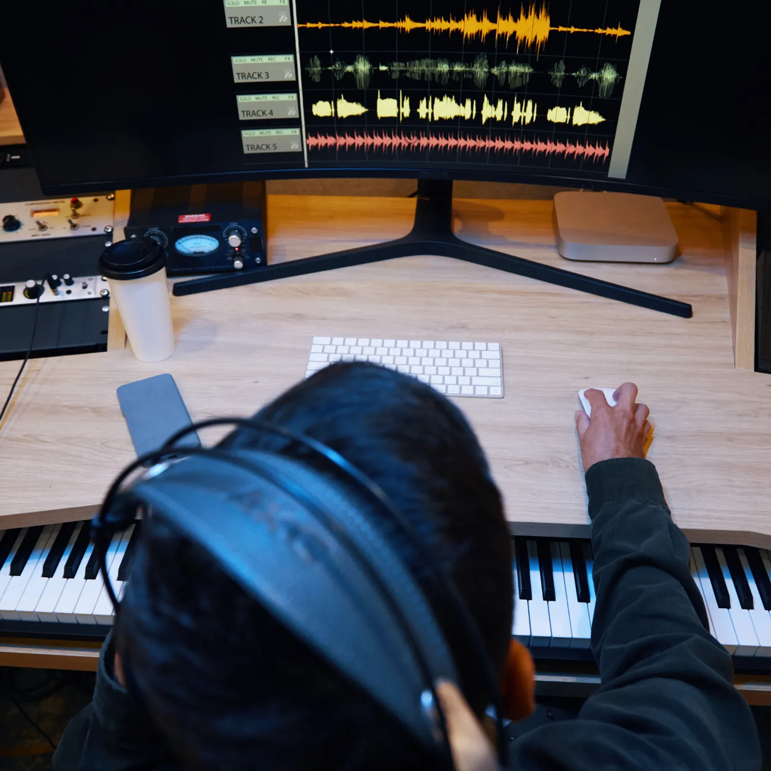 Person wearing headphones working on music editing software visible on a large curved monitor above a keyboard and desk with mouse, keyboard, phone, and coffee cup.