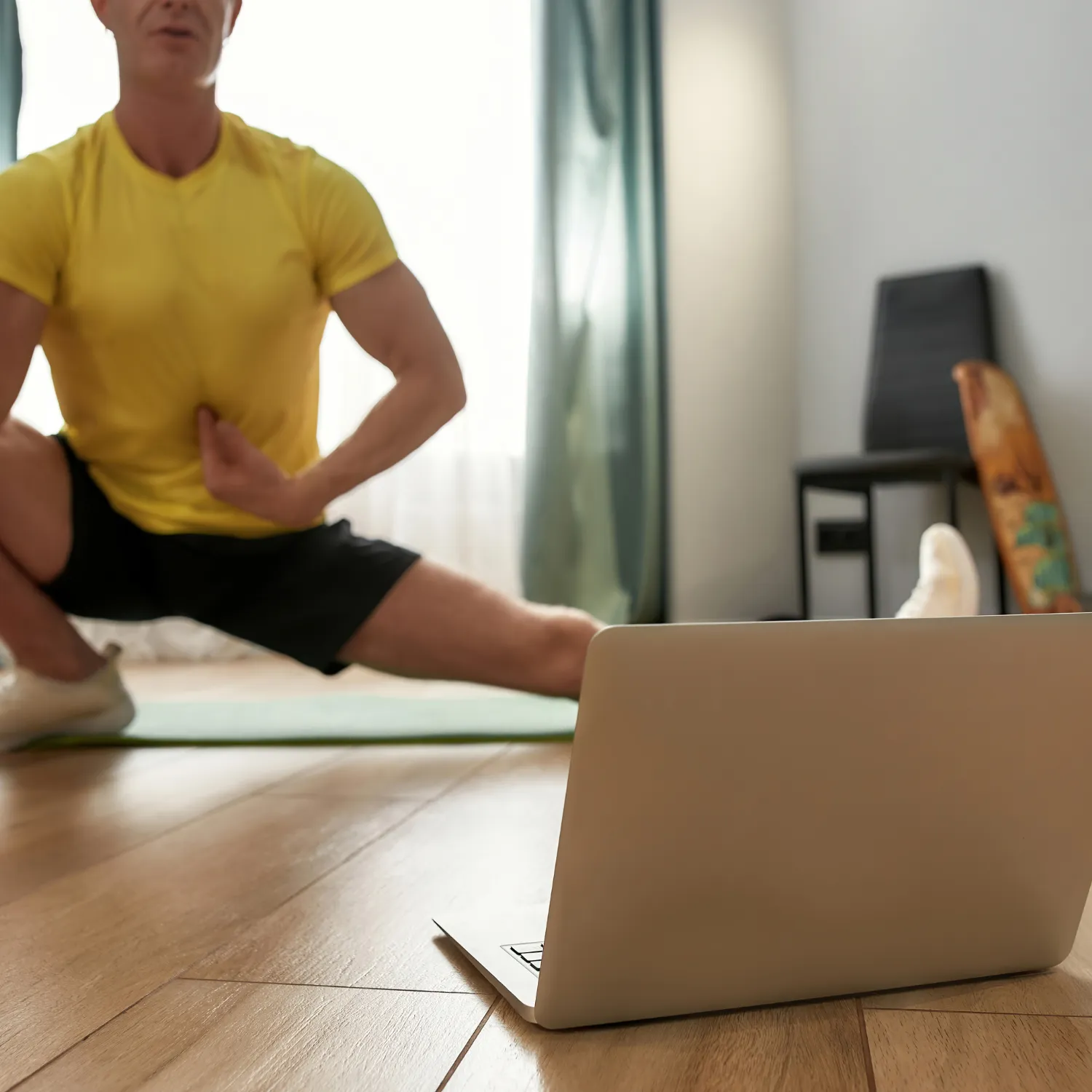 Man in yellow shirt doing a side lunge stretch on a yoga mat following a workout on a laptop at home.
