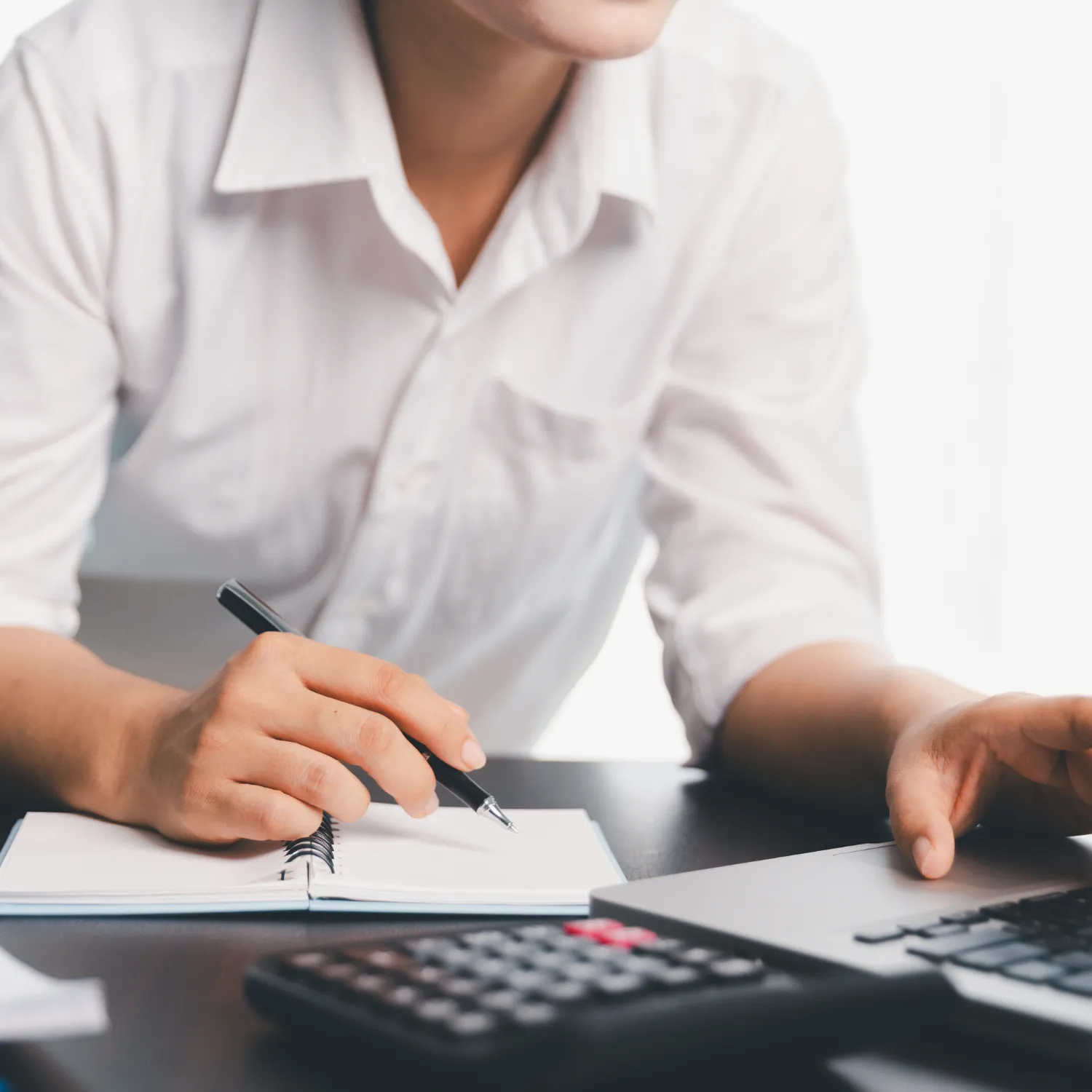 Person in white shirt writing in a notebook with a pen, working at a desk with a calculator and laptop.