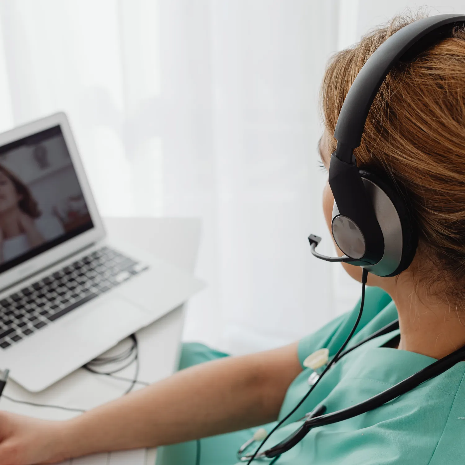 Medical professional wearing green scrubs and a headset, conducting a video call on a laptop.