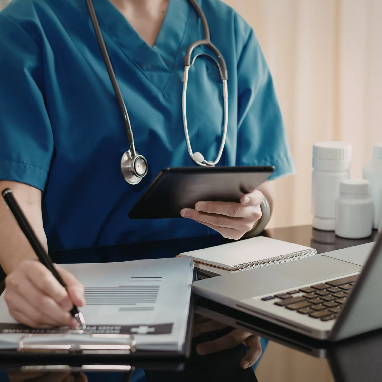 Healthcare professional in blue scrubs using a tablet and writing on a clipboard with medicine bottles and a laptop on the desk.