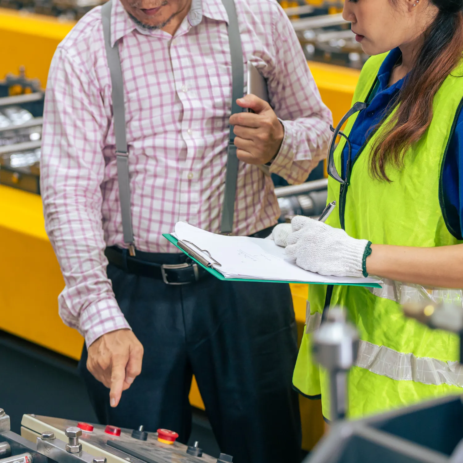 Two factory workers reviewing a clipboard with the man pointing at control buttons on a machine.