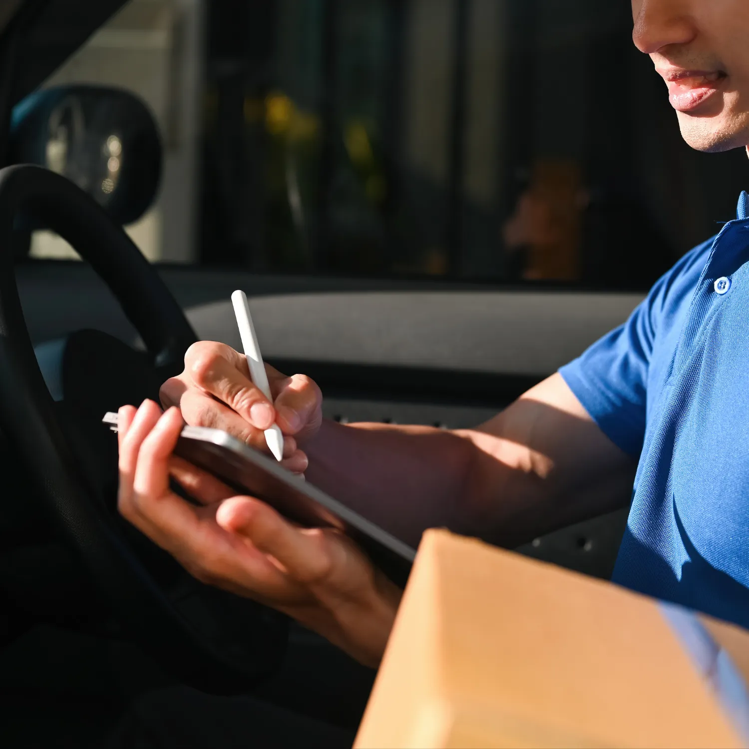 Delivery person in blue shirt inside a vehicle signing on a tablet while holding a package.