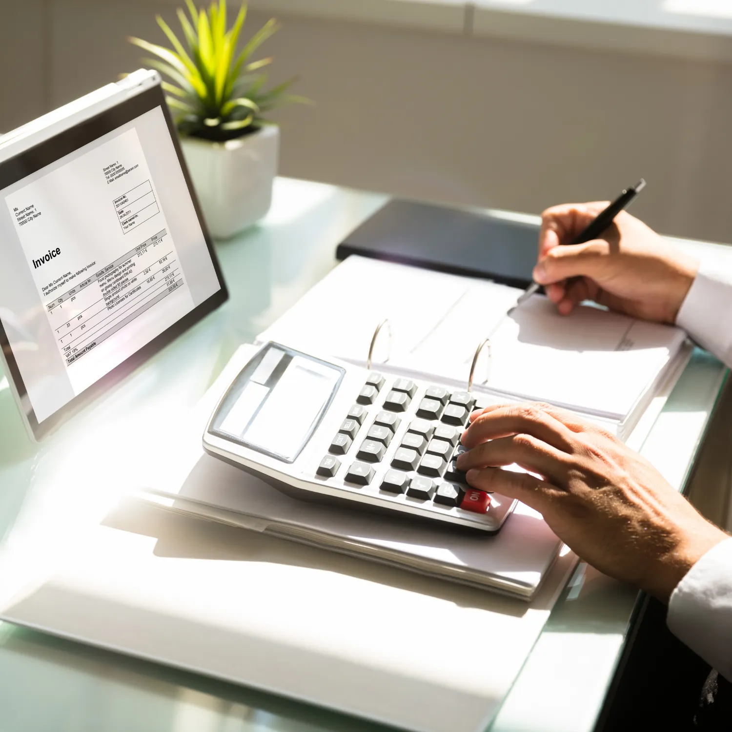 Person using a calculator and writing in a notebook beside a monitor displaying an invoice, with a small plant in the background.