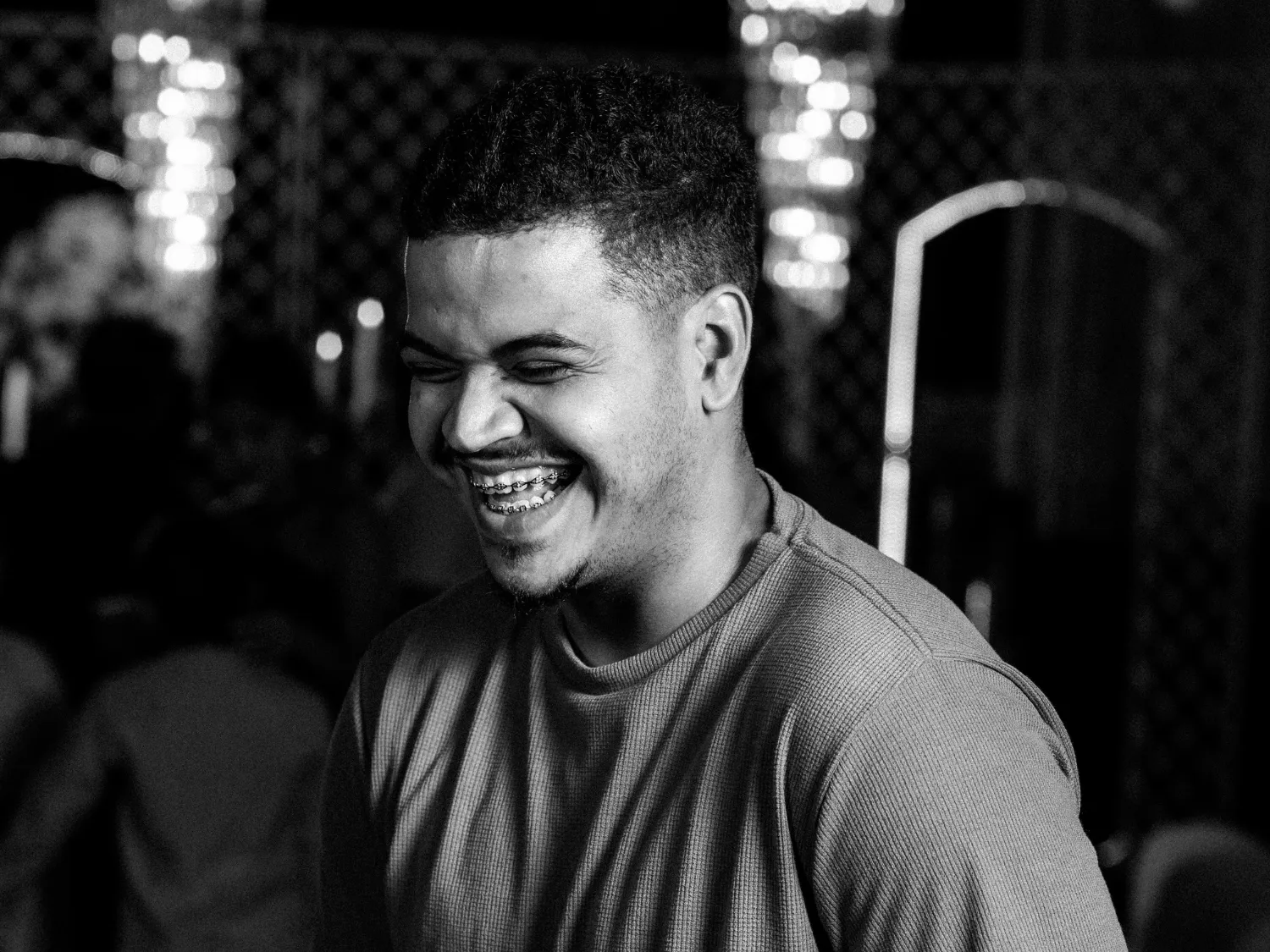 Man with braces smiling and looking down, wearing a ribbed long-sleeve shirt, in a dimly lit indoor setting.