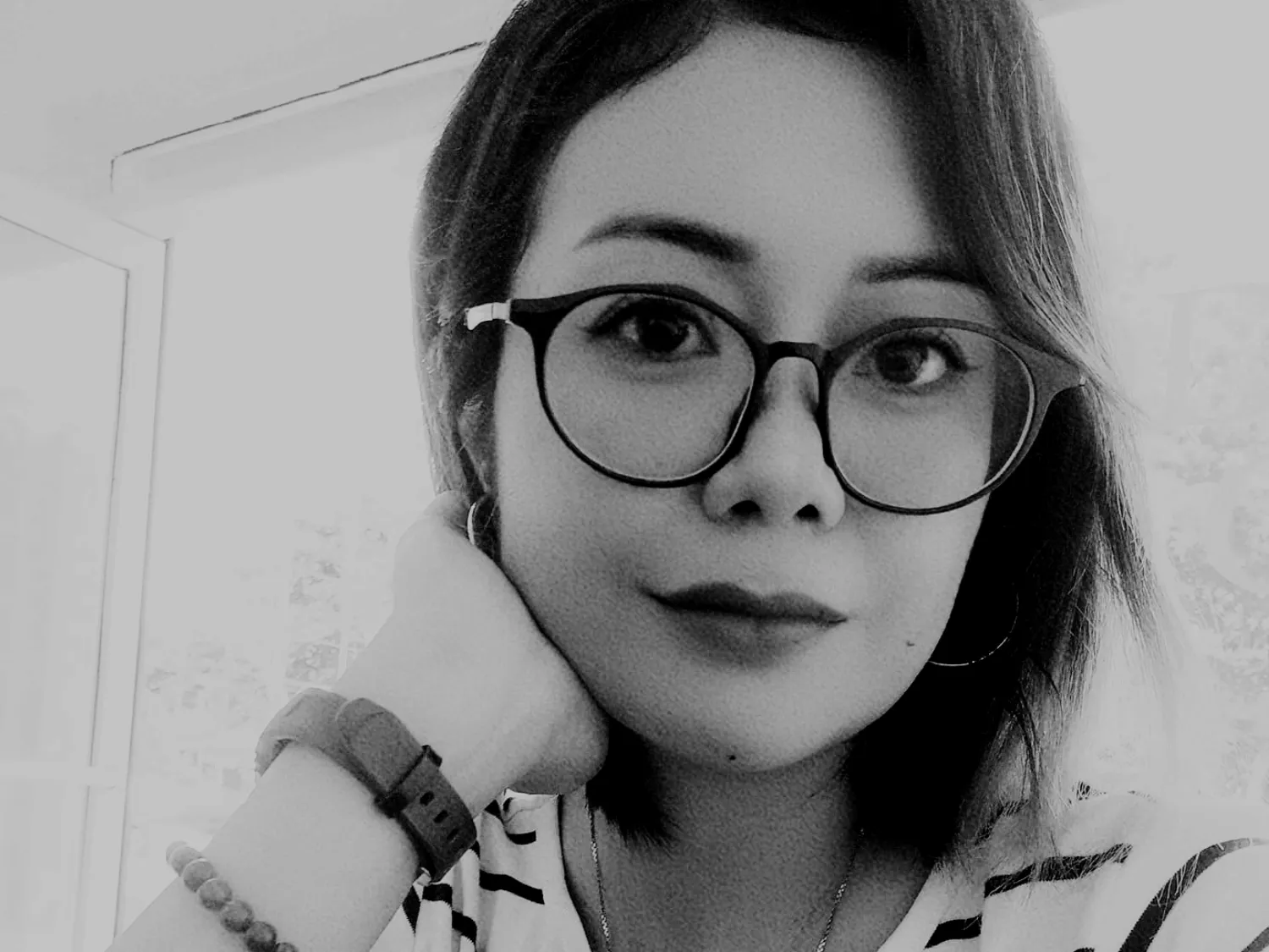 Close-up of a woman with short hair wearing large round glasses, hoop earrings, and a striped shirt, resting her hand on her cheek.