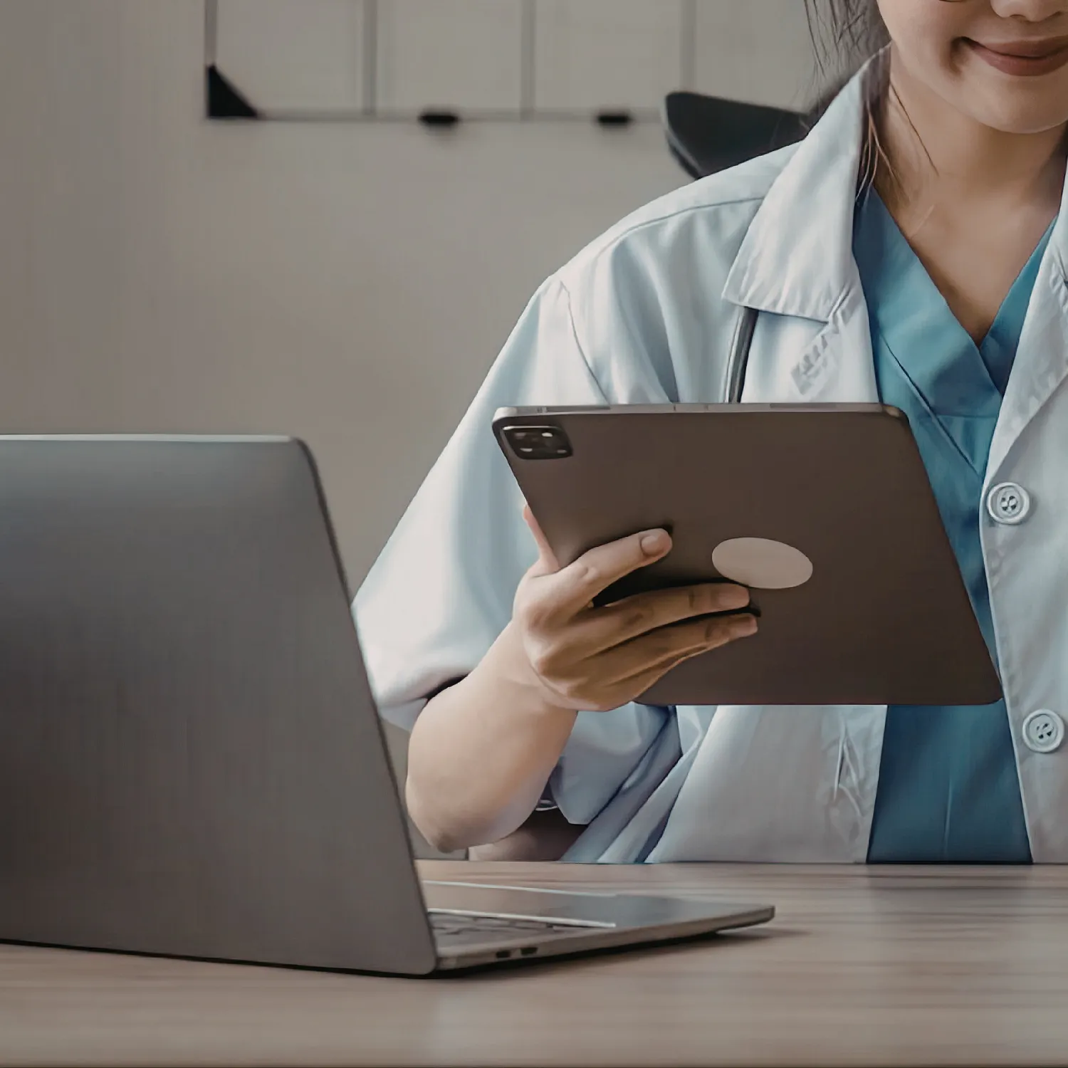 Healthcare professional in scrubs and white coat holding a tablet, sitting at a desk with a laptop.
