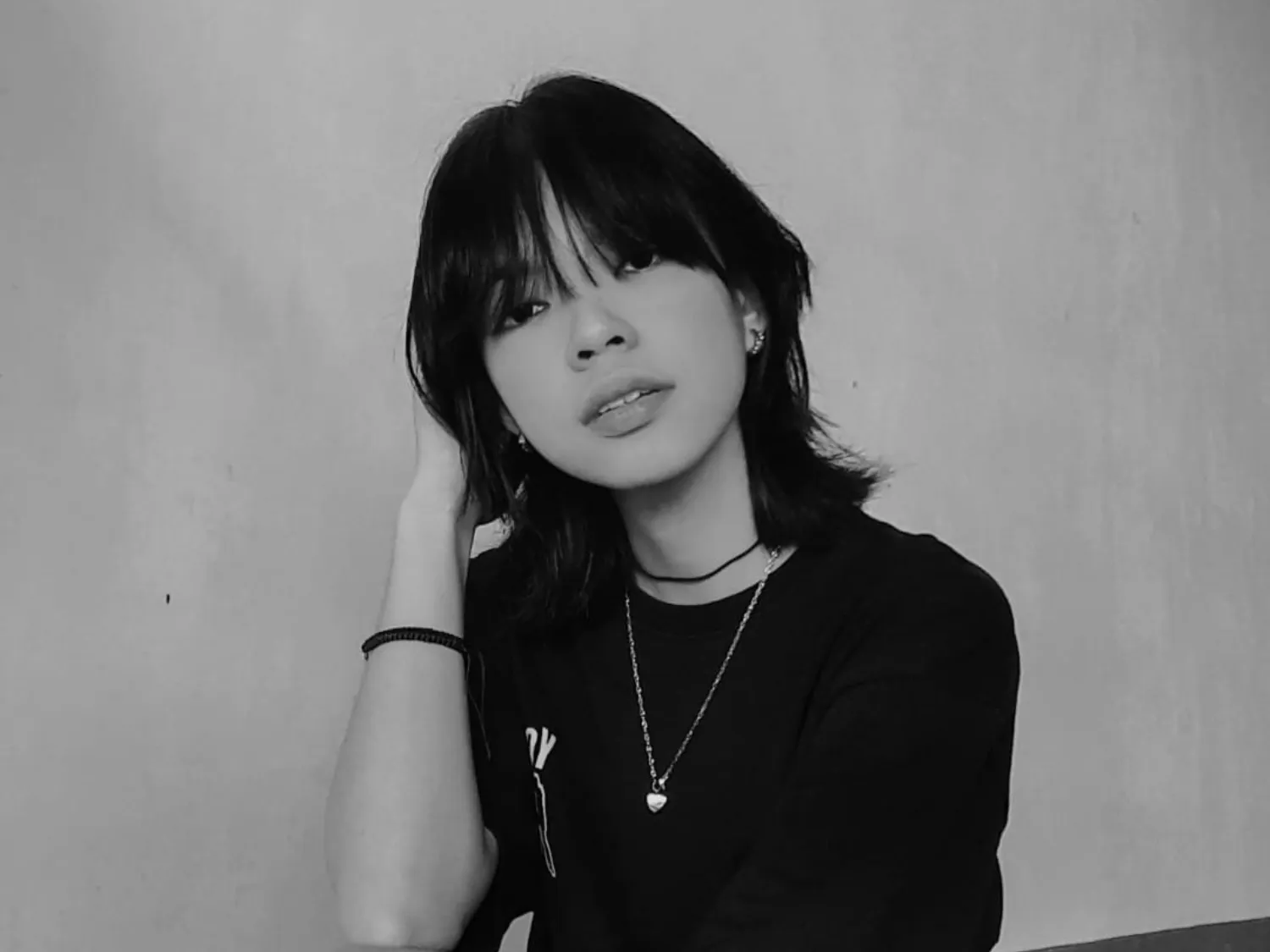 Black and white photo of a young woman with shoulder-length hair resting her head on her hand, wearing a necklace and a dark shirt.