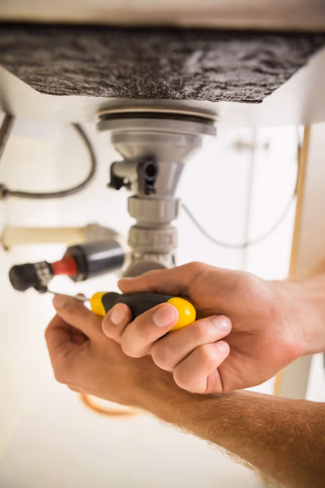 Handen van een loodgieter die een schroevendraaier gebruiken om leidingen onder een gootsteen te repareren.