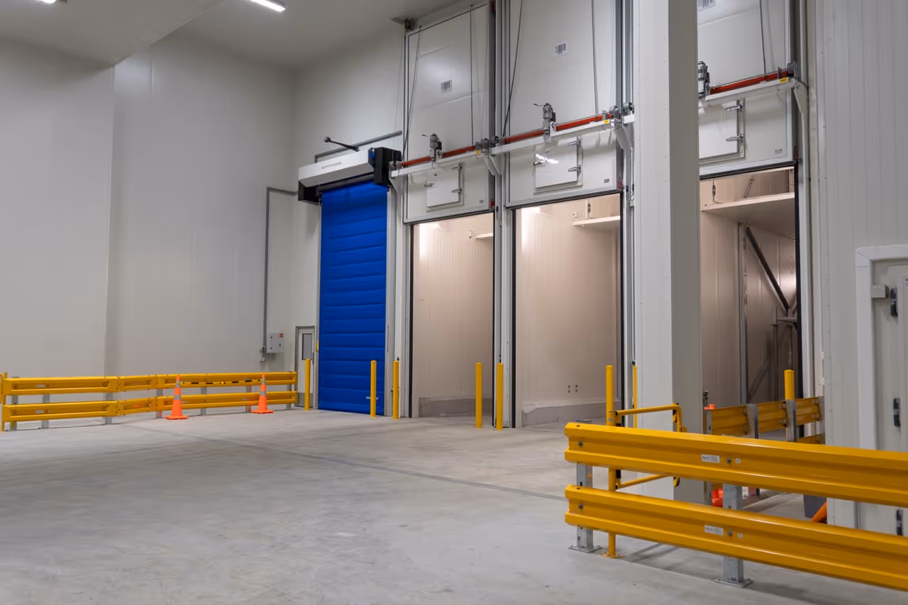Interior of a warehouse with large open loading dock doors, yellow safety barriers, and a blue overhead door.