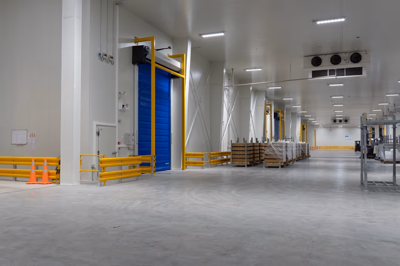 Spacious industrial warehouse interior with blue roller doors, yellow guardrails, stacked pallets, and concrete floor.