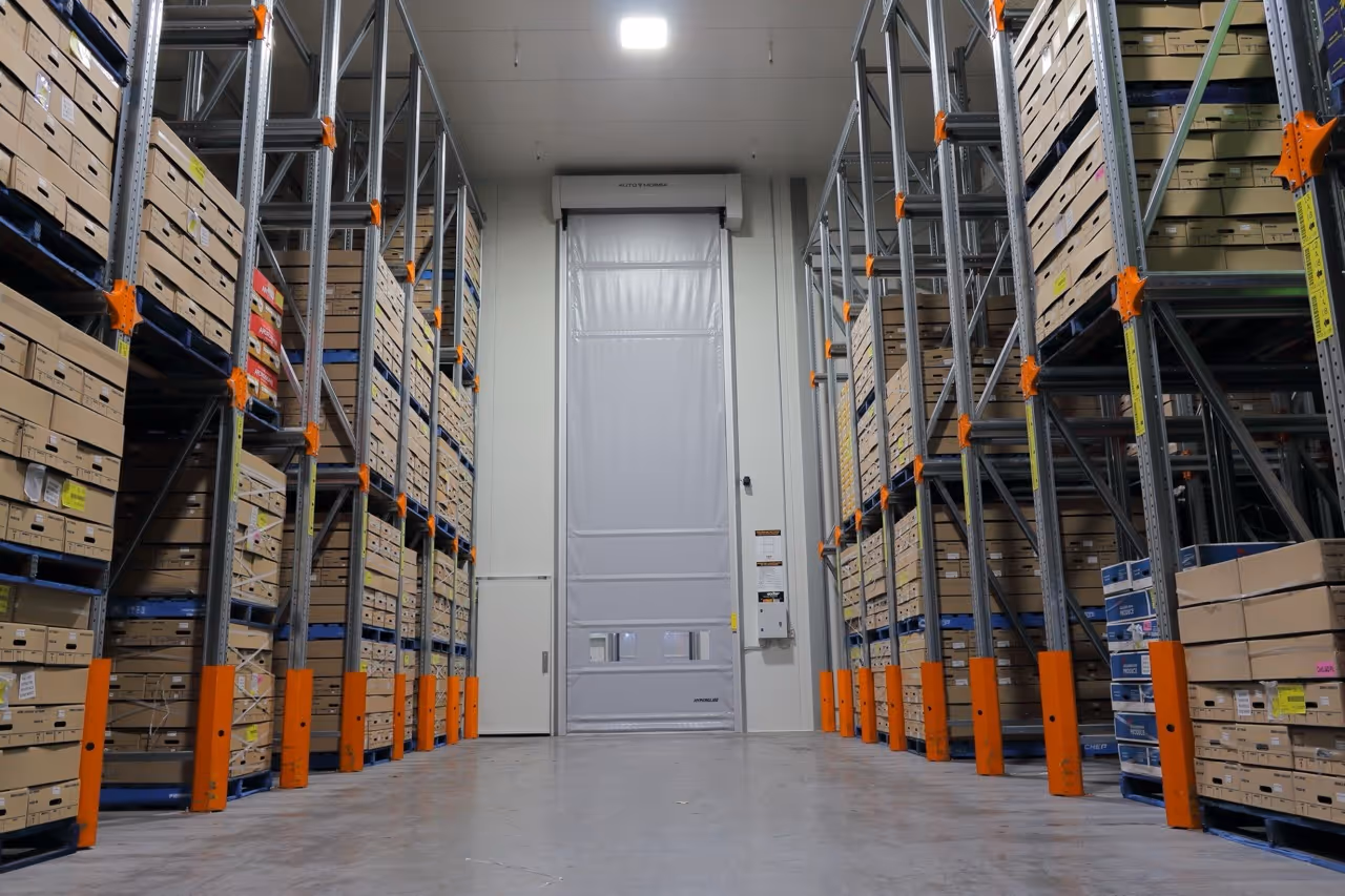 Interior of a warehouse aisle with tall metal shelves stocked with cardboard boxes and a closed white industrial door at the end.