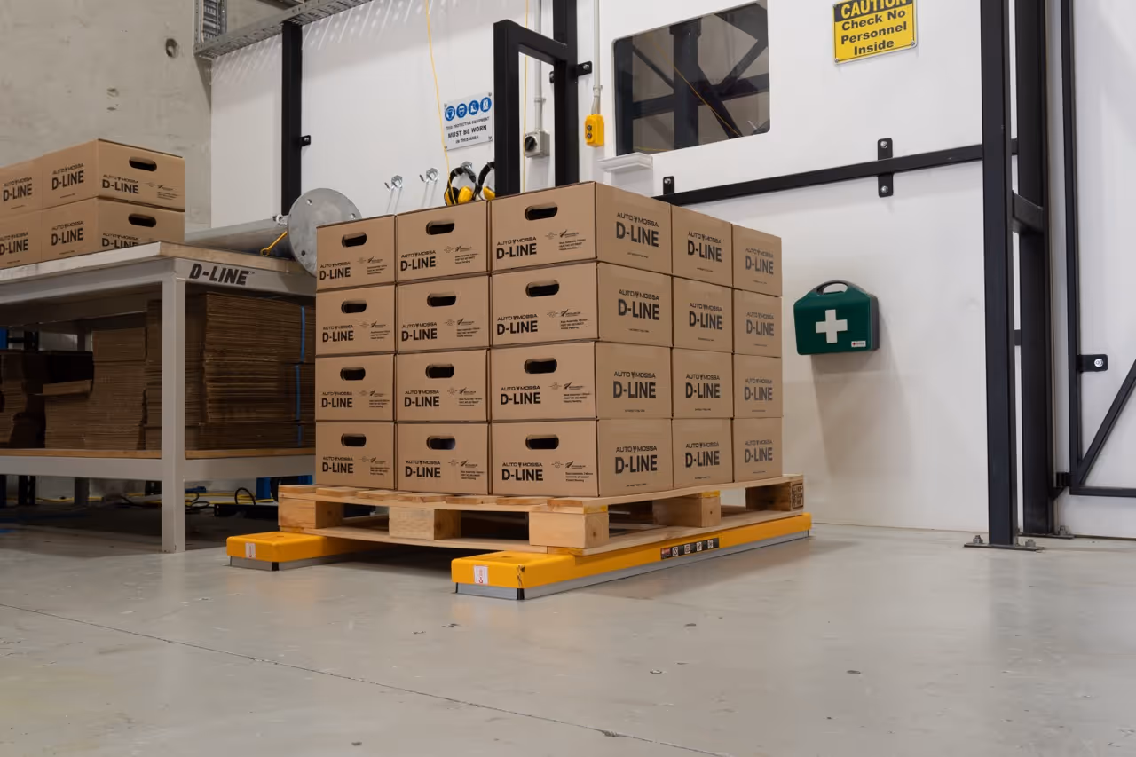 Stack of AUTOYMOOBSA D-LINE cardboard boxes on a wooden pallet in a warehouse, near a green first aid kit and caution sign.