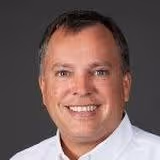 Smiling middle-aged man with short dark hair wearing a white shirt against a grey background.