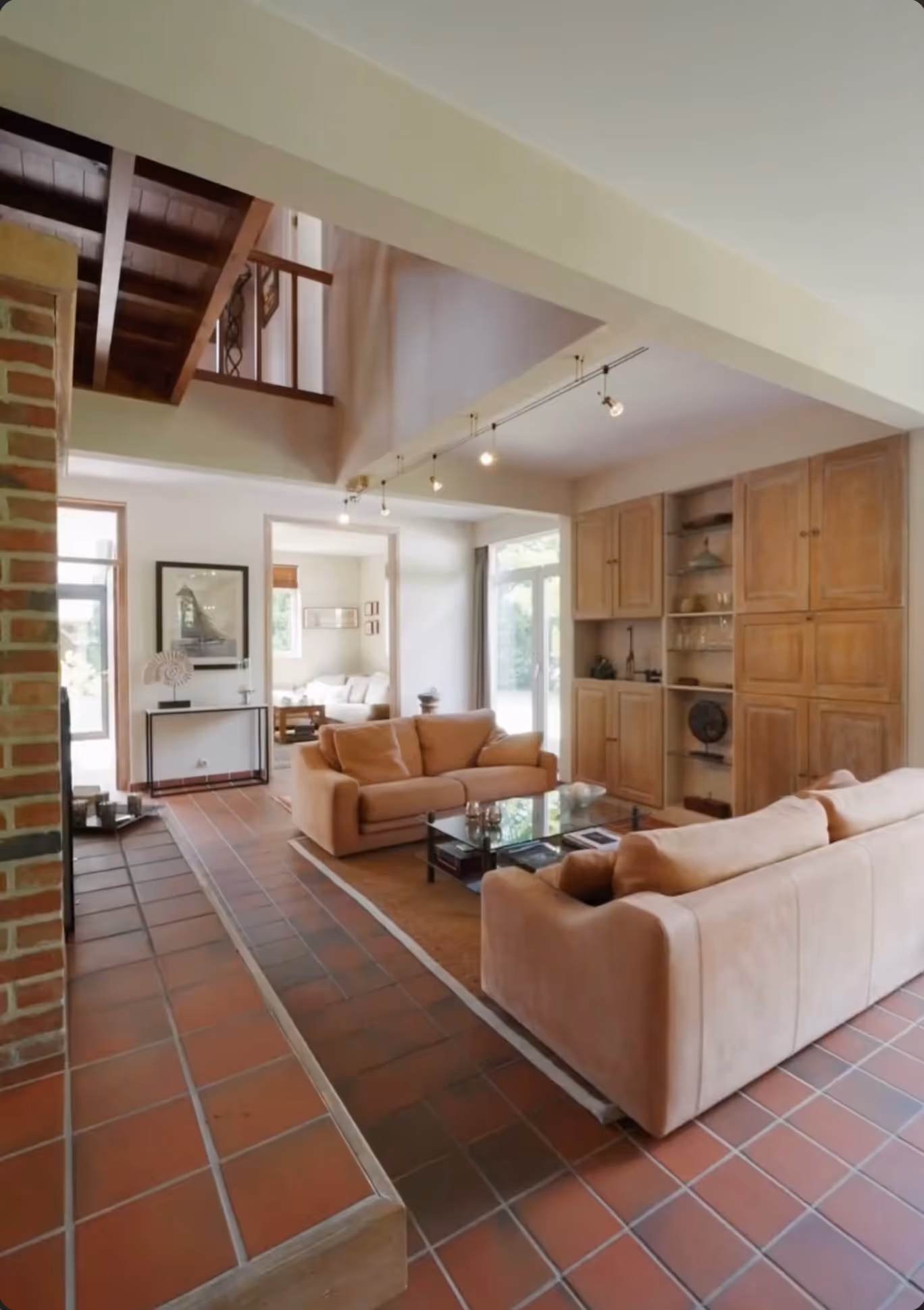 Woonkamer met terracotta vloertegels, twee beige banken rond een glazen salontafel, ingebouwde houten kasten en zicht op een aangrenzende zithoek.