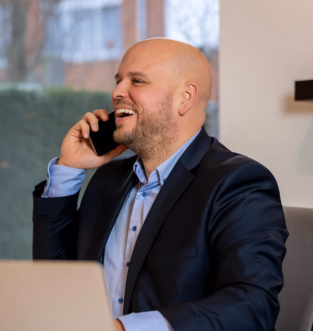 Lachende man in pak belt op mobiele telefoon terwijl hij aan een bureau zit.