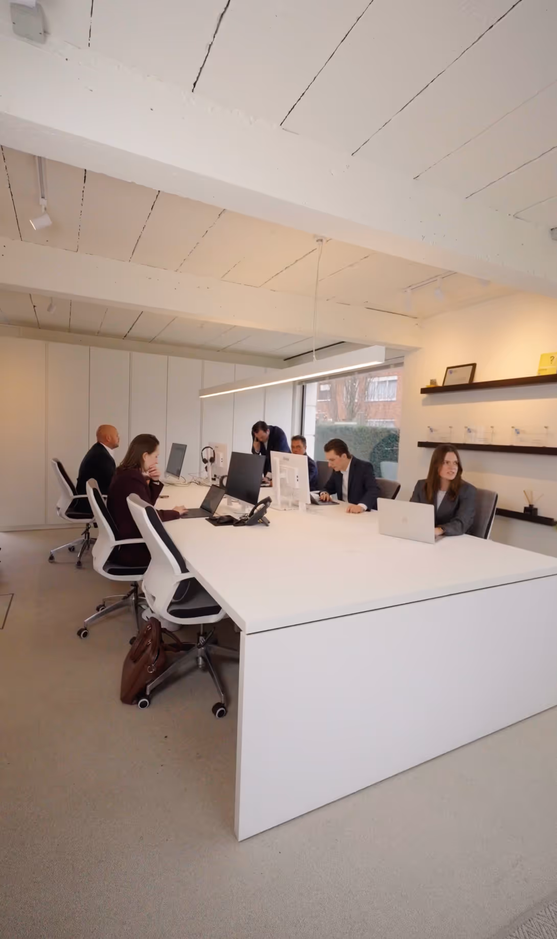 Vijf mensen werken aan laptops en computers aan een grote witte kantoorruimte met een raam en planken aan de muur.