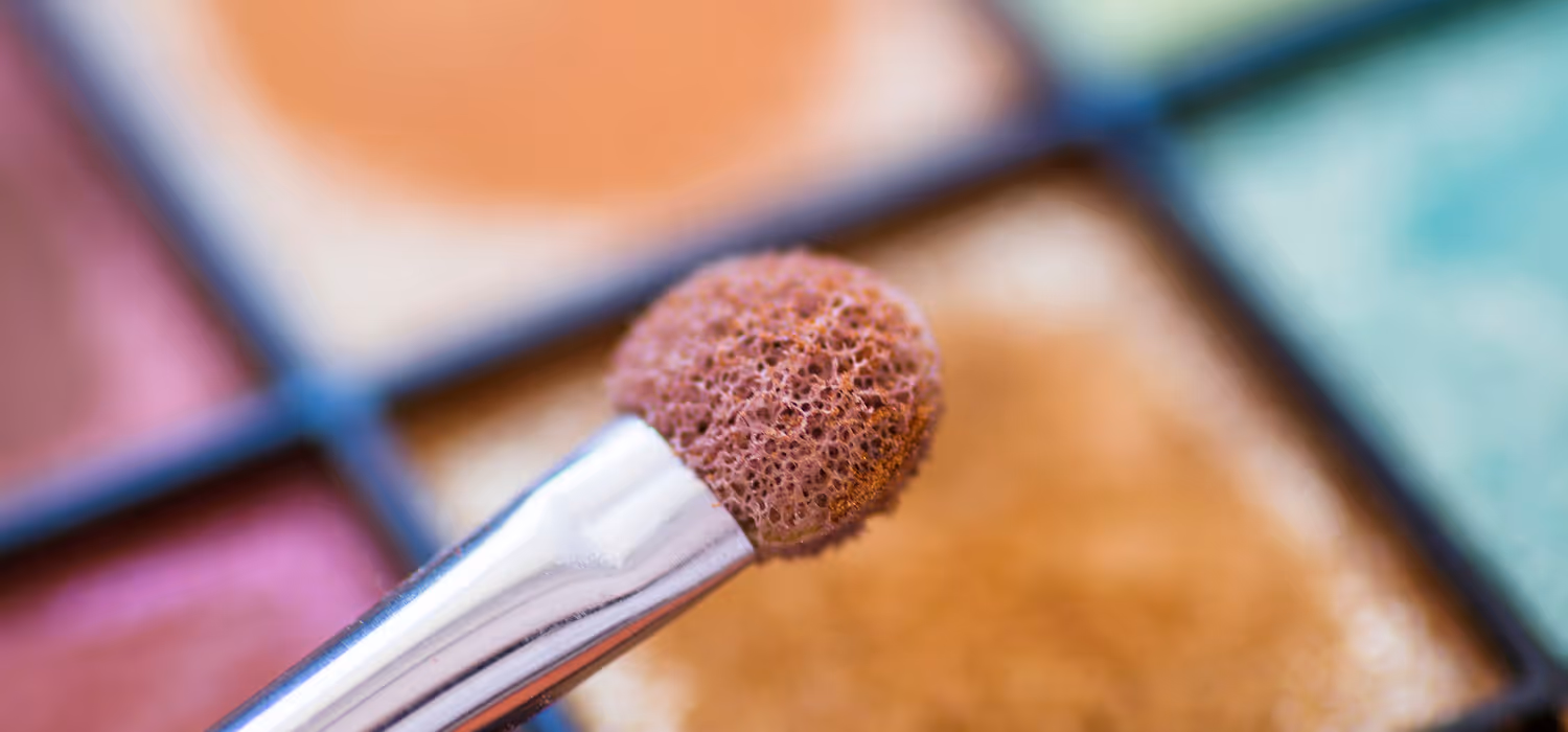 Ein Pinsel mit braunem Lidschatten vor einer Palette mit buntem Lidschatten.