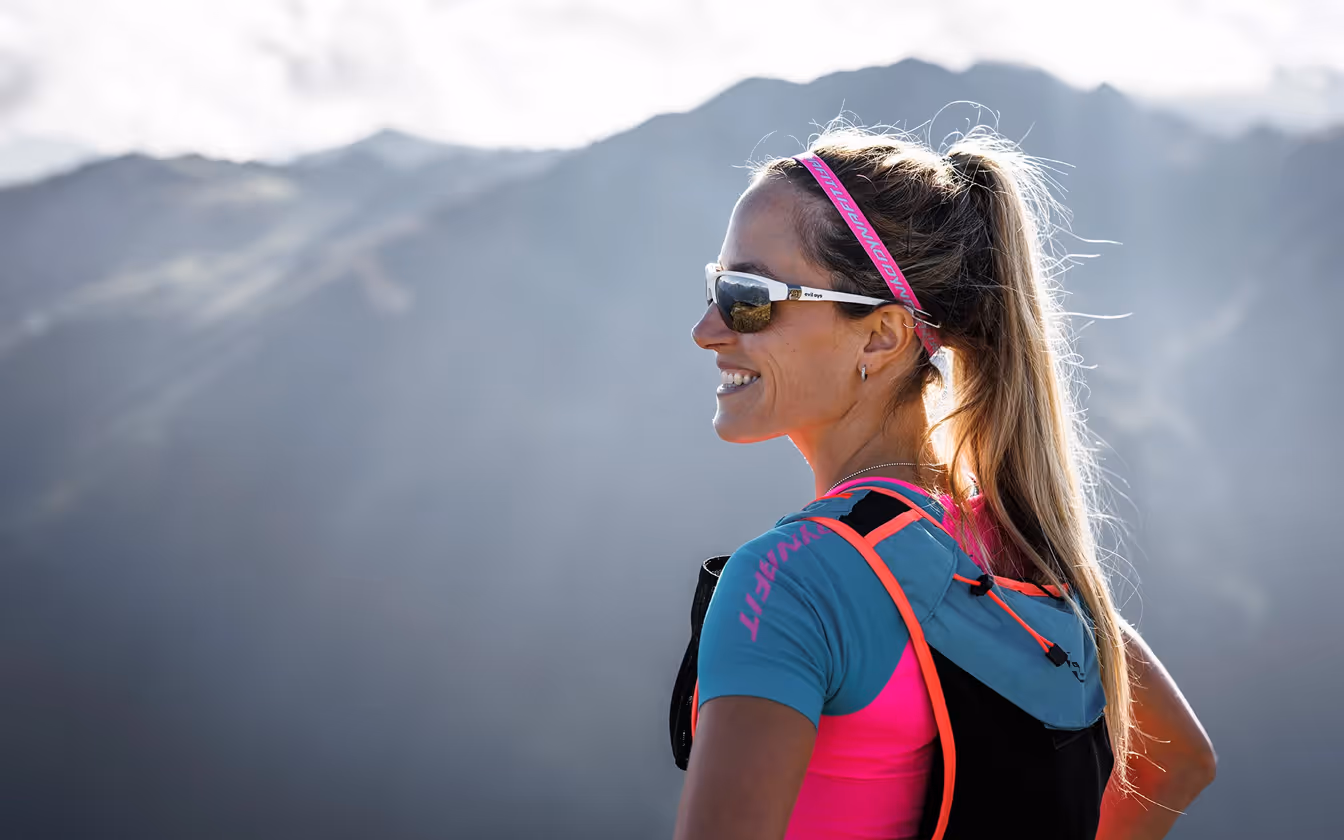 Frau mit Pferdeschwanz, Sportbrille und Sportbekleidung mit Rucksack, die lächelnd vor Berglandschaft steht.