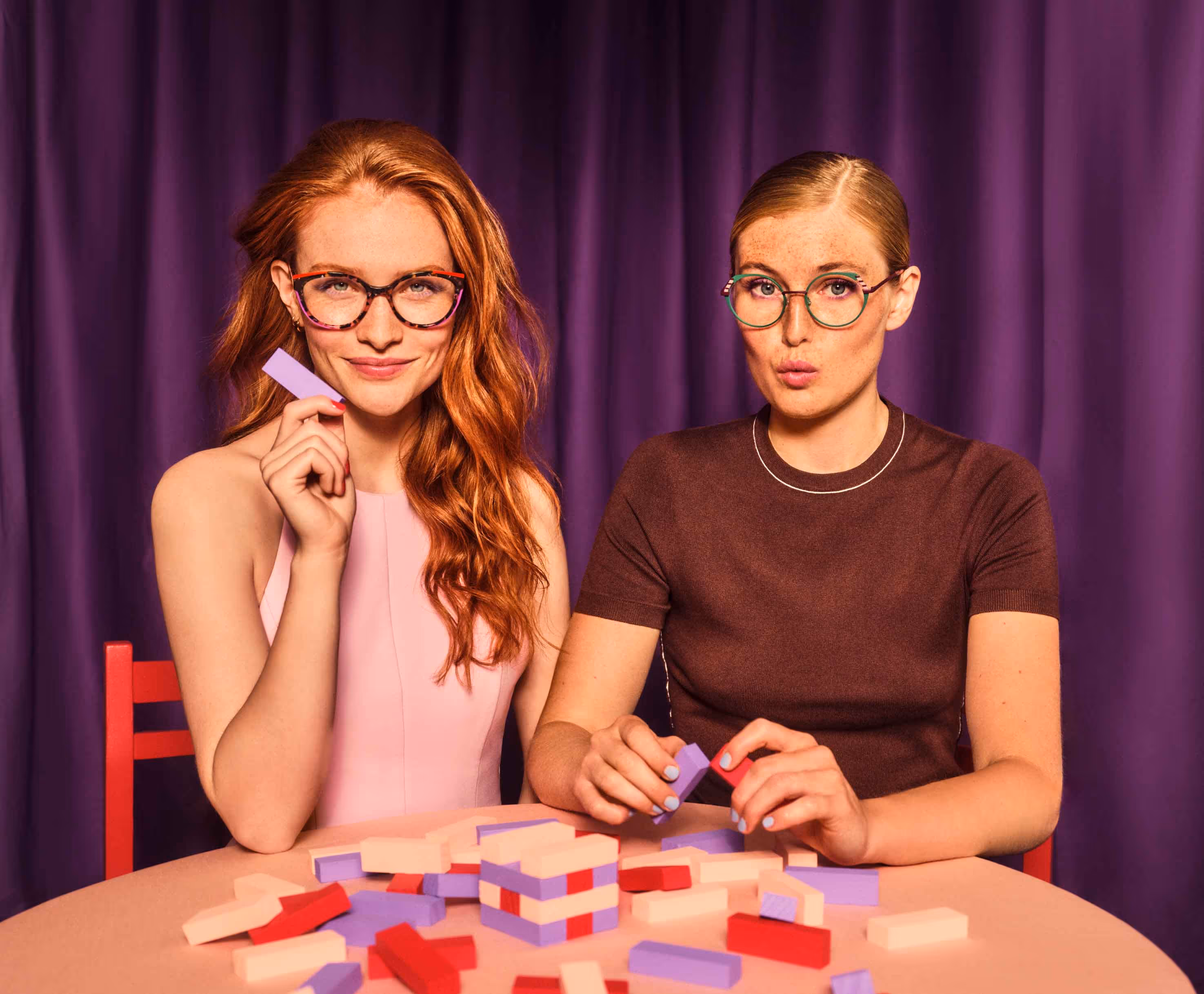 Zwei Frauen mit Brillen sitzen an einem Tisch mit bunten Spielsteinen vor einem lila Vorhang.