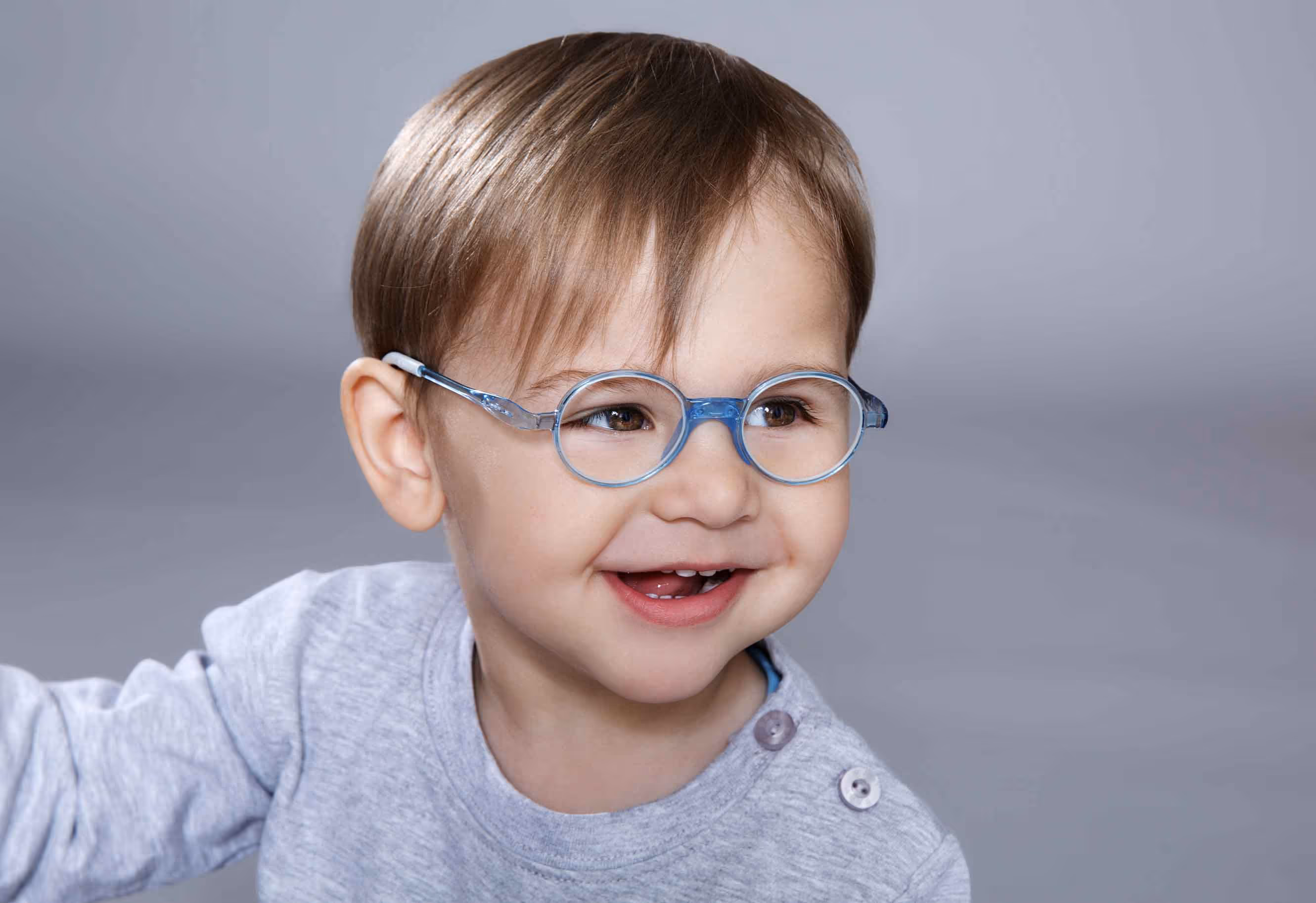 Kleines Kind mit kurzen braunen Haaren und blauen runden Brillengestellen lächelt freundlich.