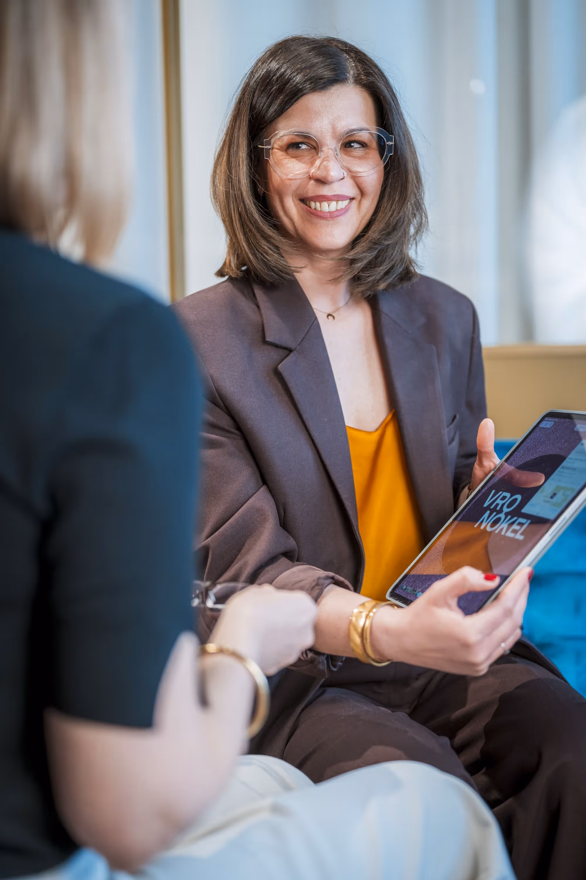 Zwei Frauen in professioneller Umgebung, eine lächelnd mit Tablet, die andere unscharf im Vordergrund.