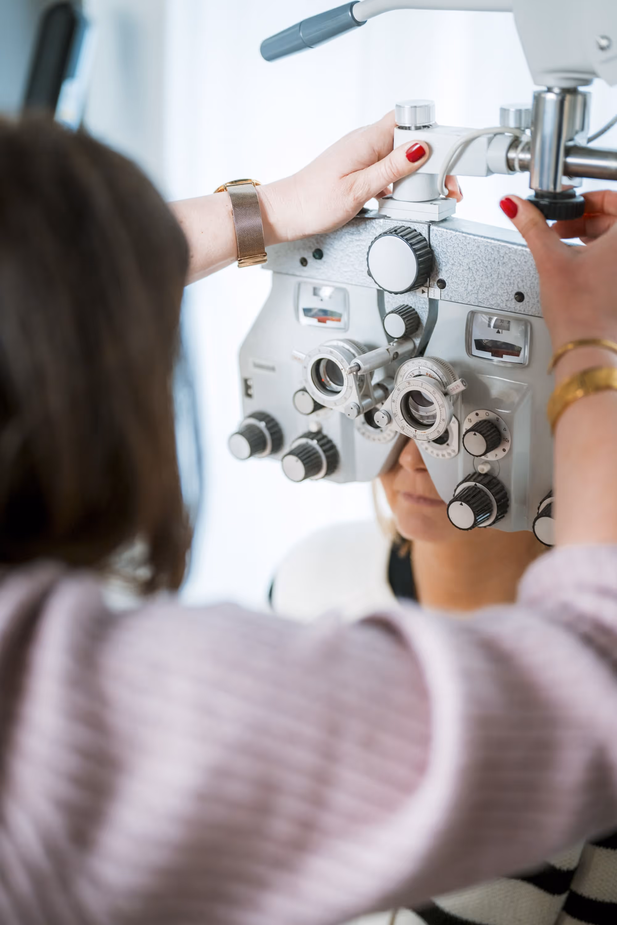 Augenuntersuchung mit einem Phoropter, bei der eine Person durch das Gerät blickt, während eine Optikerin das Gerät einstellt.