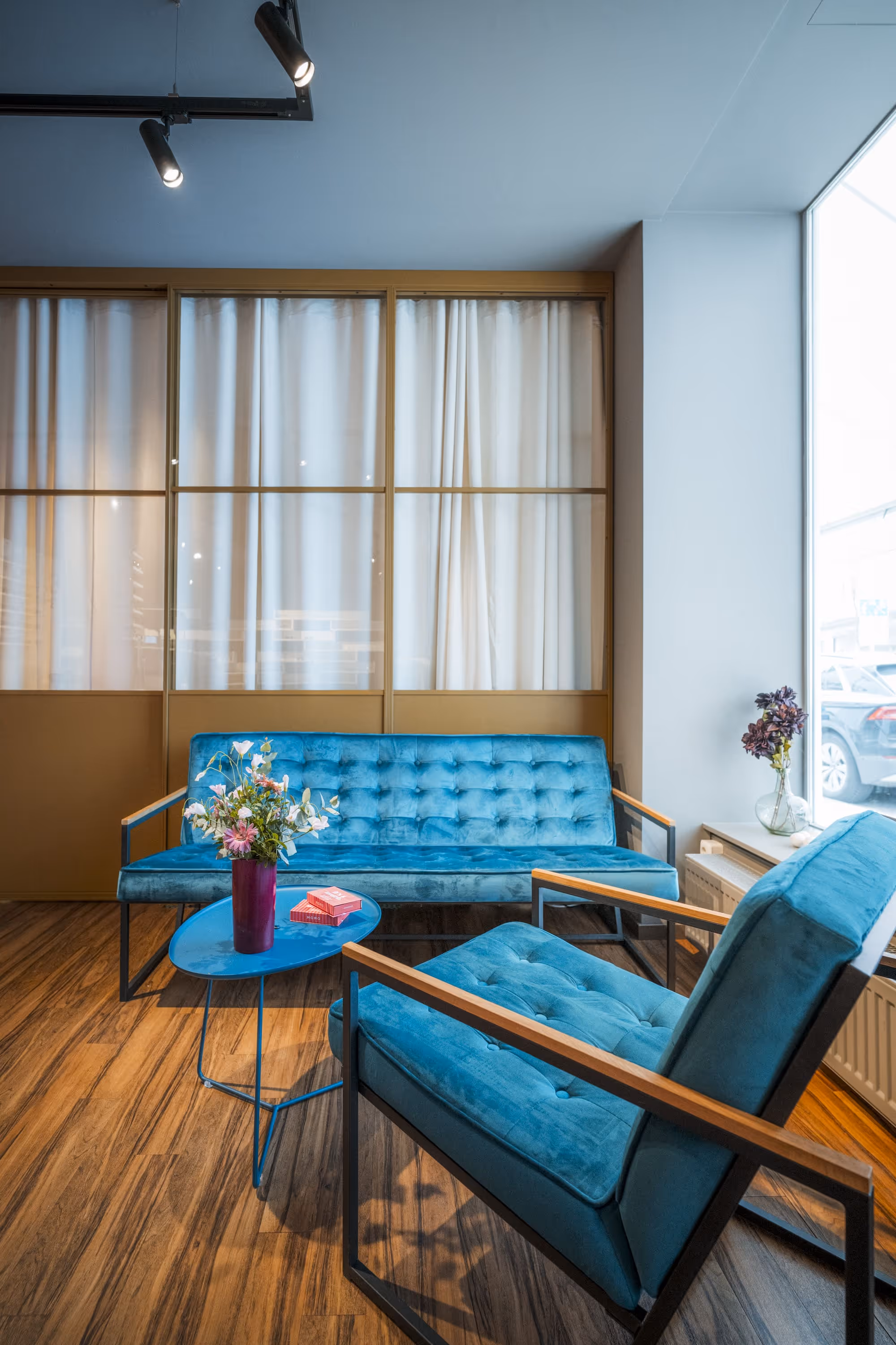 Modern sitting area with a blue velvet sofa and armchair around a small blue coffee table with a flower vase on wooden flooring.