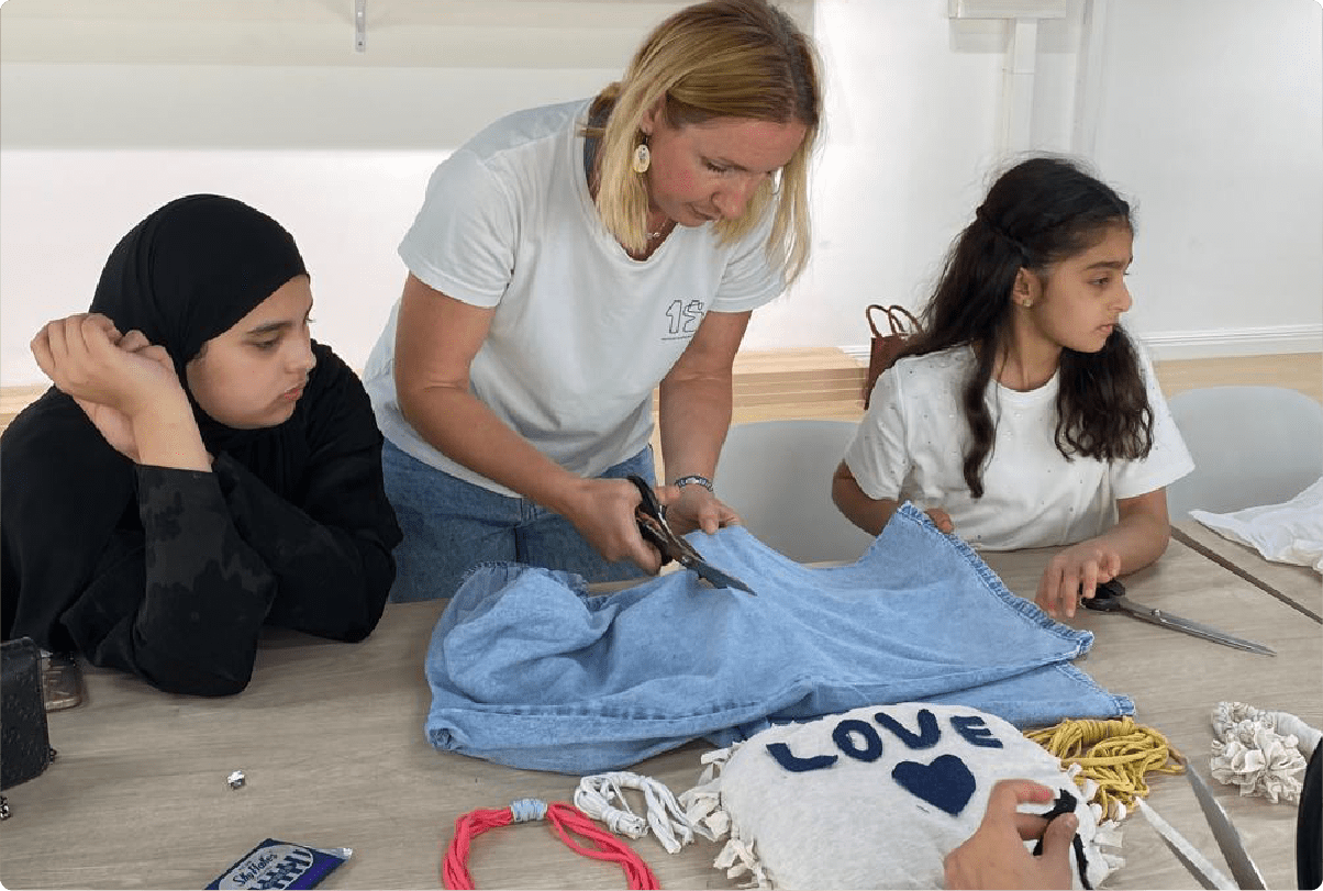 Woman helping two girls cut a pair of blue jeans at a crafting table with various materials, including a fabric piece with the word 'LOVE' and a heart.