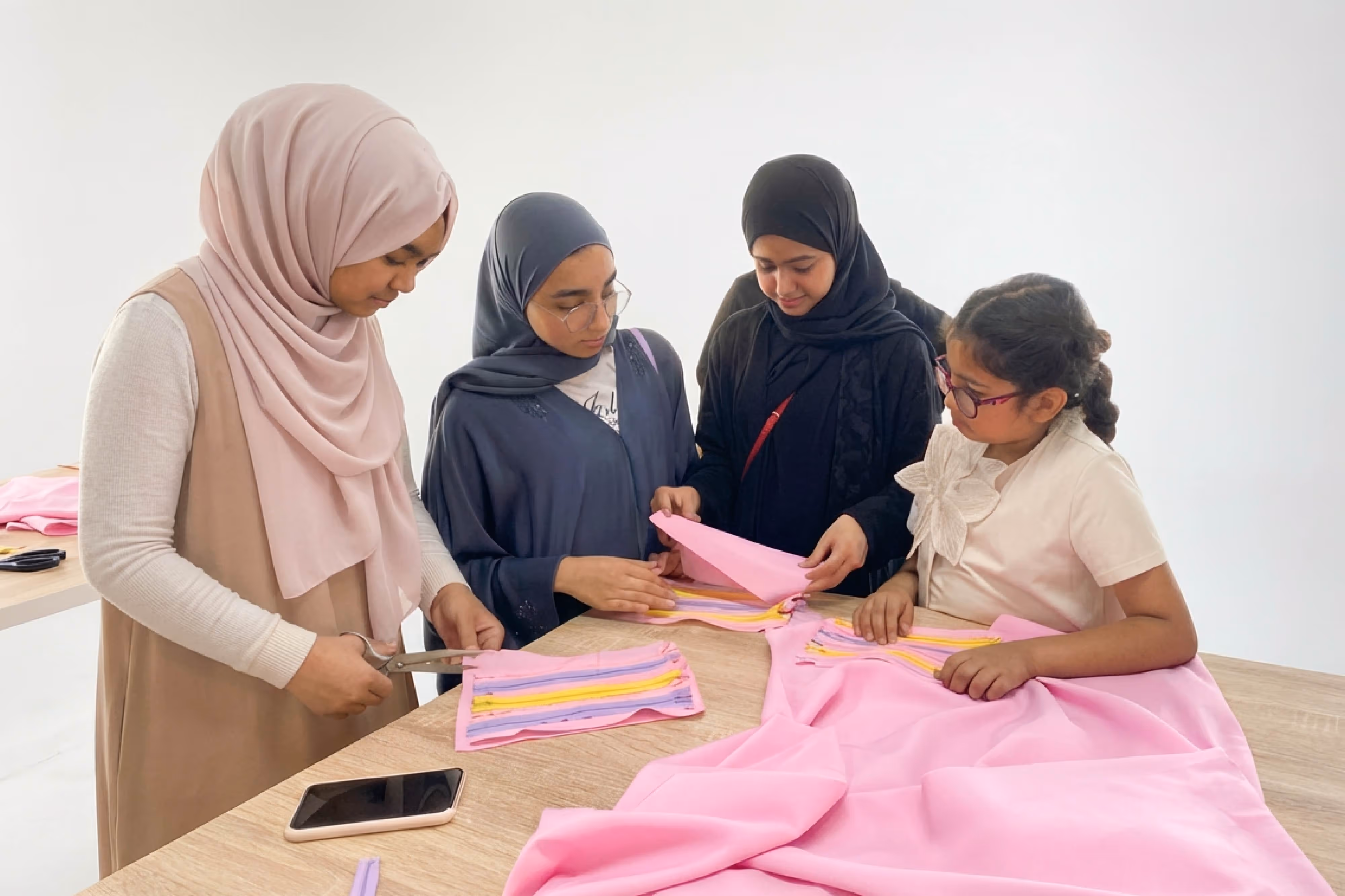Four females working together on pink fabric with colorful zippers at a table.