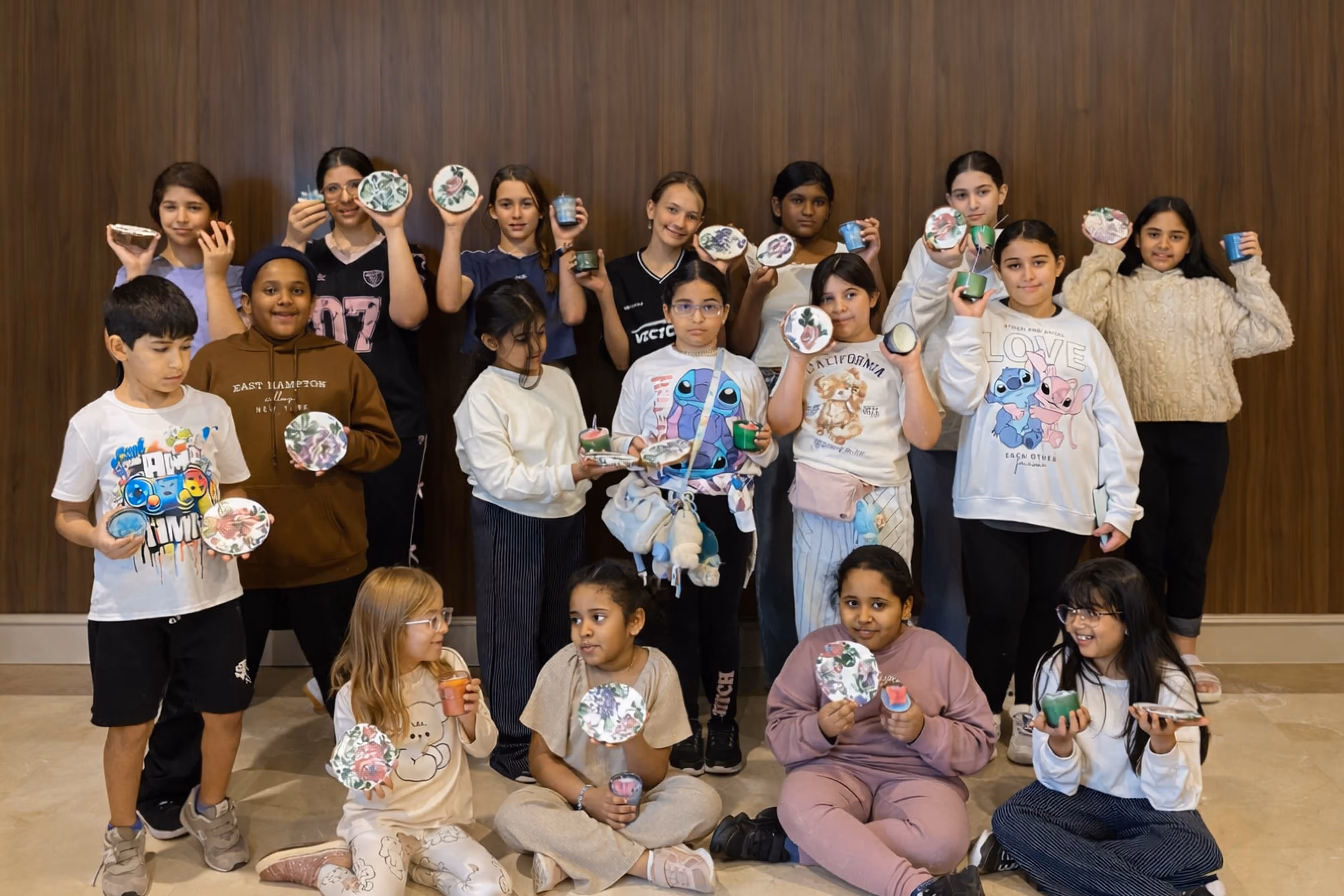 Group of children proudly displaying hand-decorated flower pots and candles in front of a wooden wall.