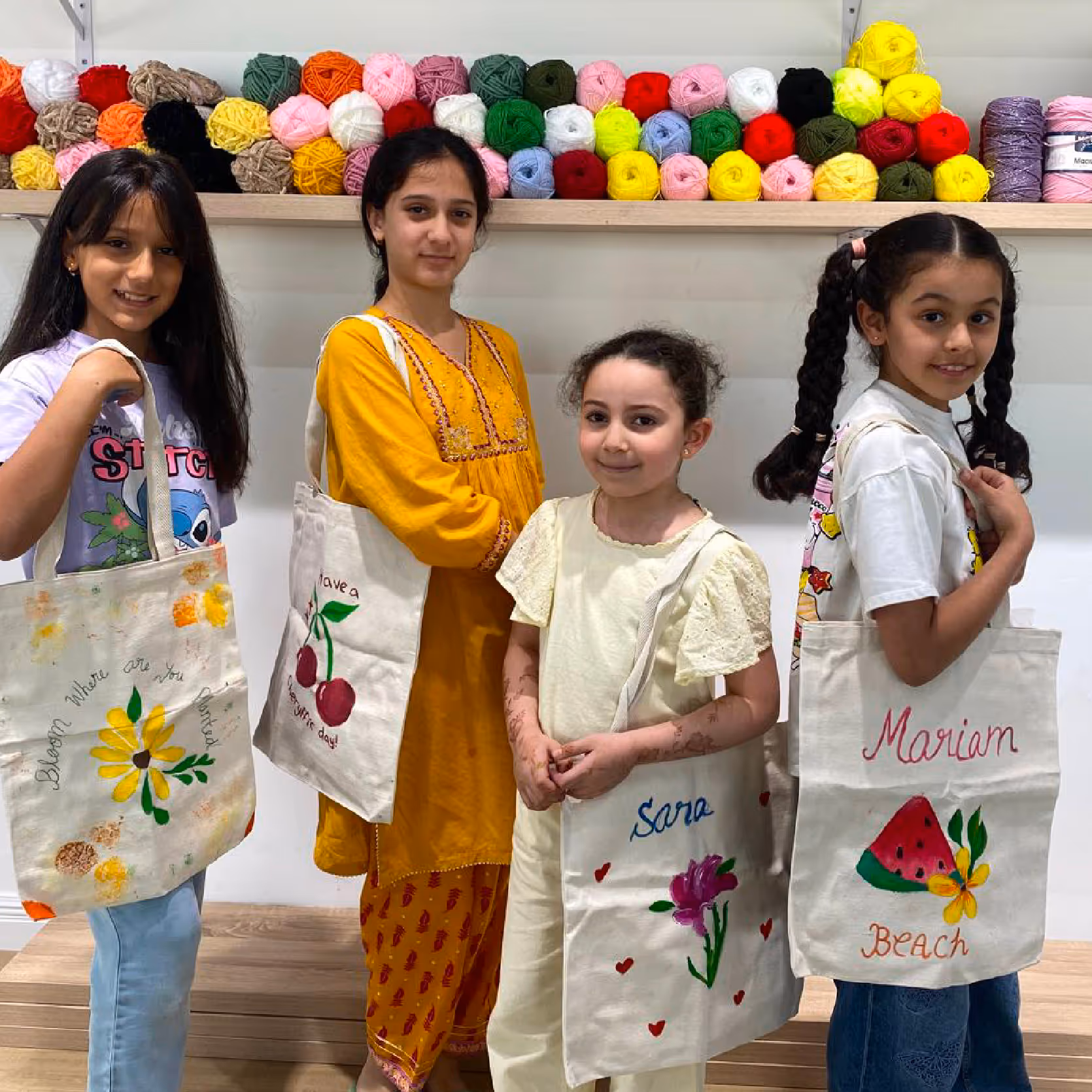 Four girls standing indoors holding decorated tote bags with floral and fruit designs, with colorful yarn balls on a shelf behind them.