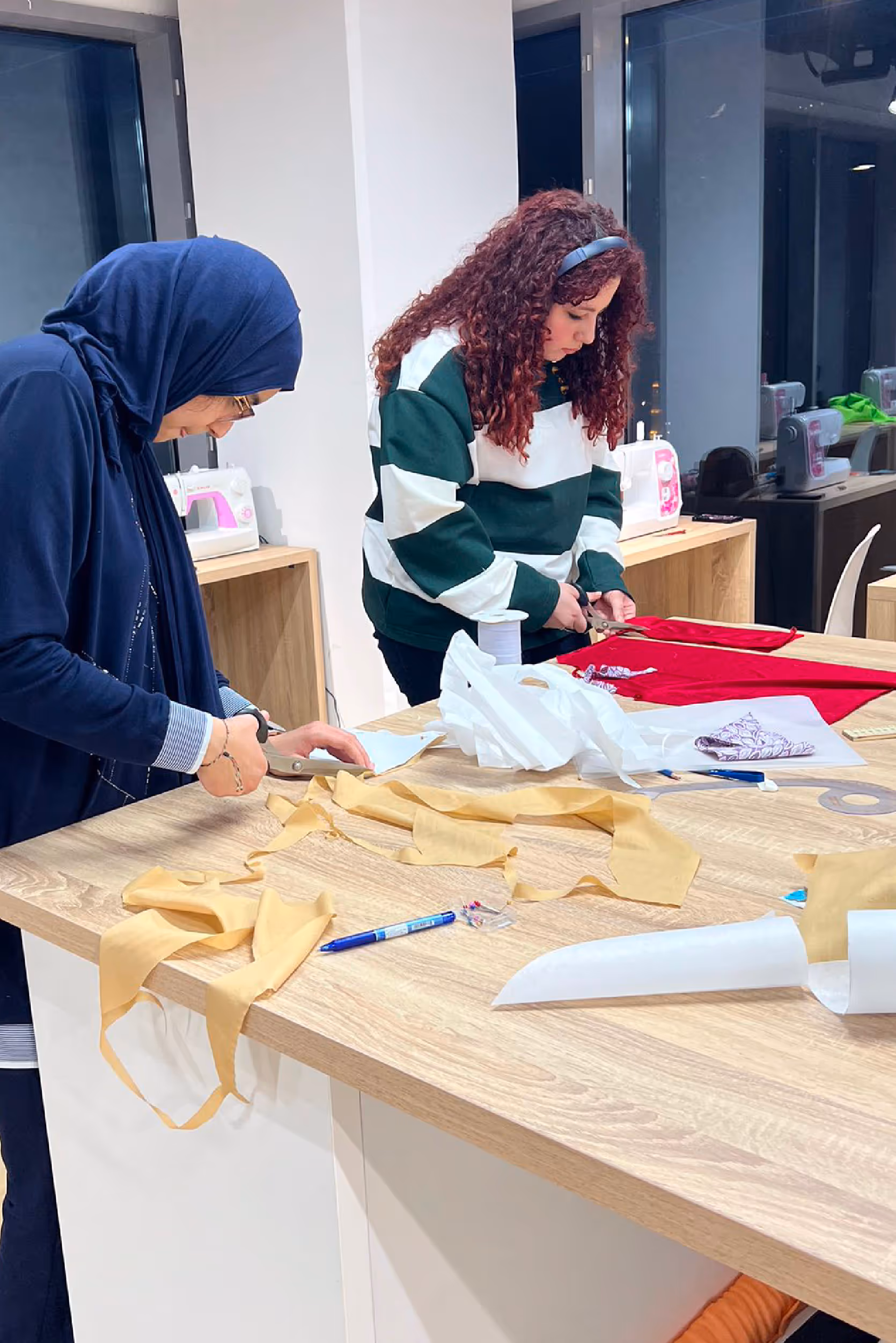 Two women cutting fabric on a wooden table in a sewing studio, with sewing machines in the background.