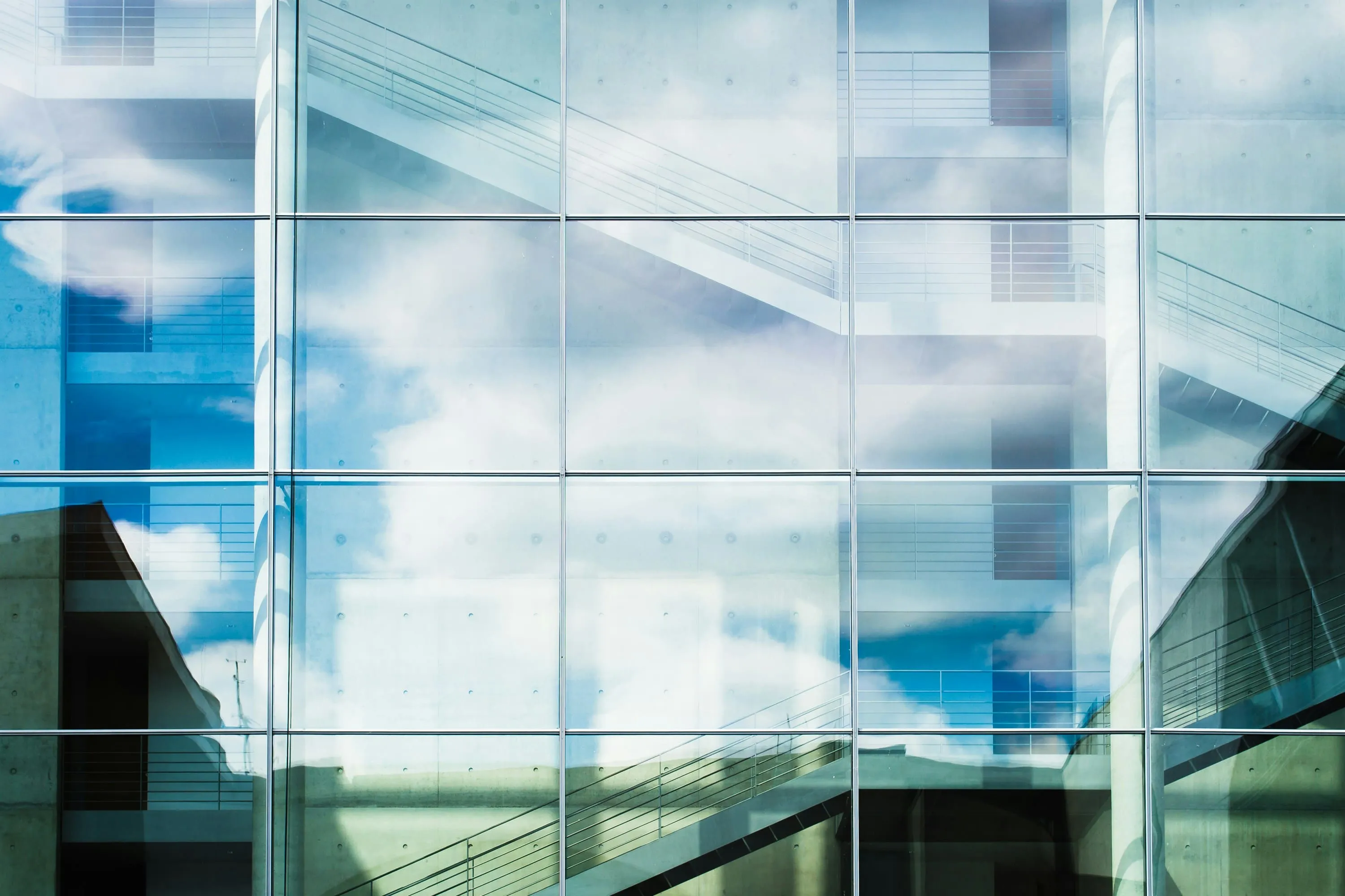 Reflection of clouds and blue sky on a glass building facade with visible staircases behind the glass.