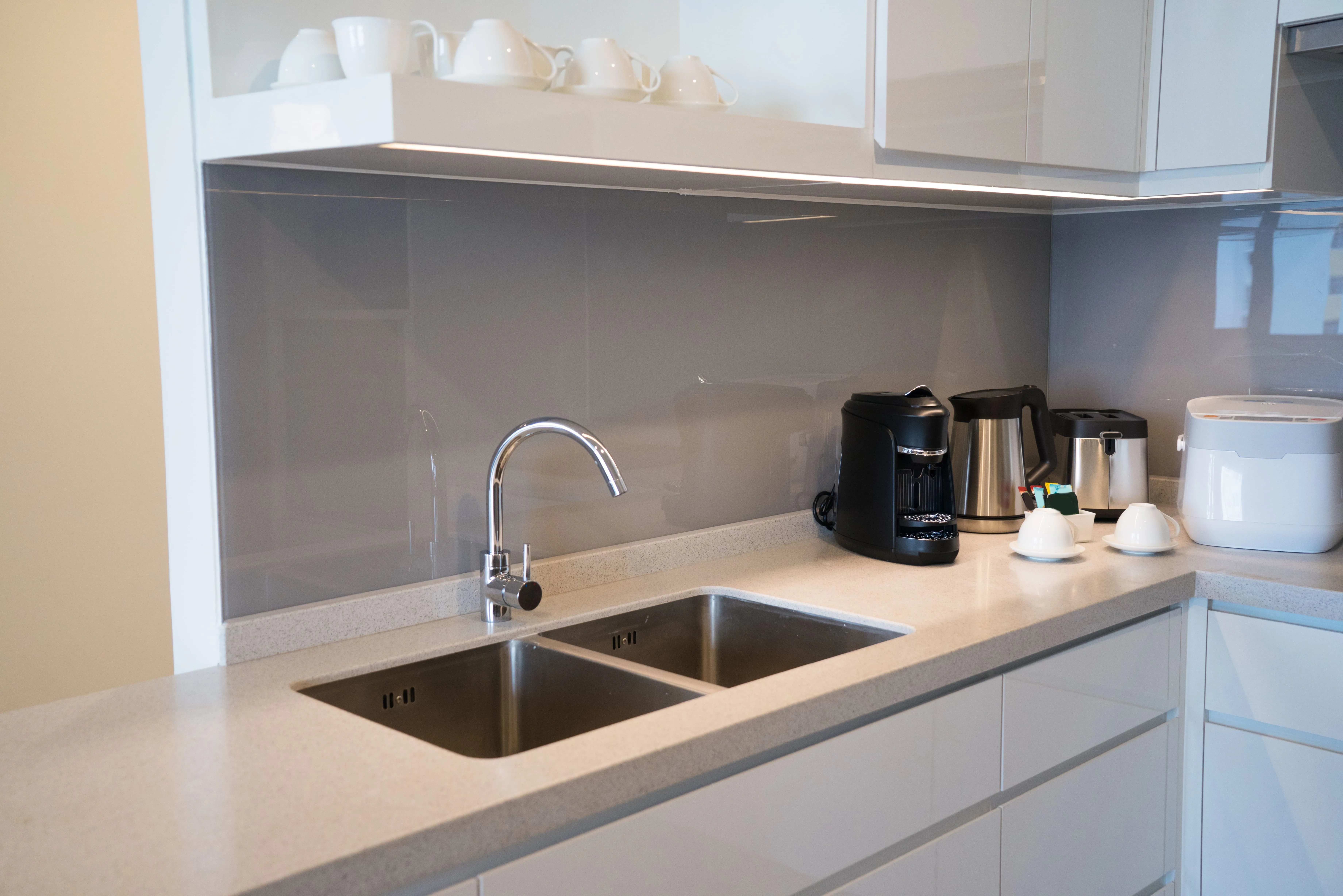 Modern kitchen countertop with double sink, chrome faucet, coffee machine, kettle, toaster, and cups against a grey splashback.